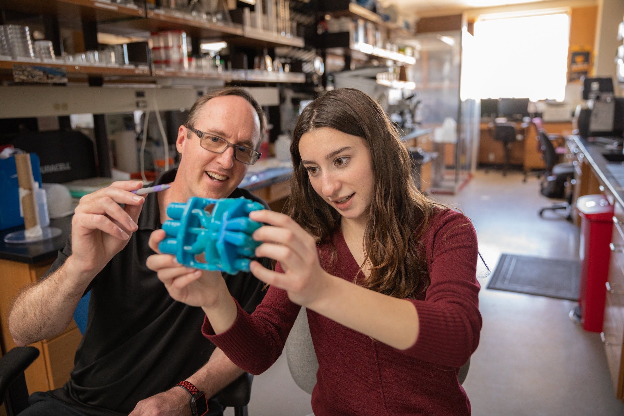 A man and a young woman examine a blue 3D-printed object together in a laboratory setting.