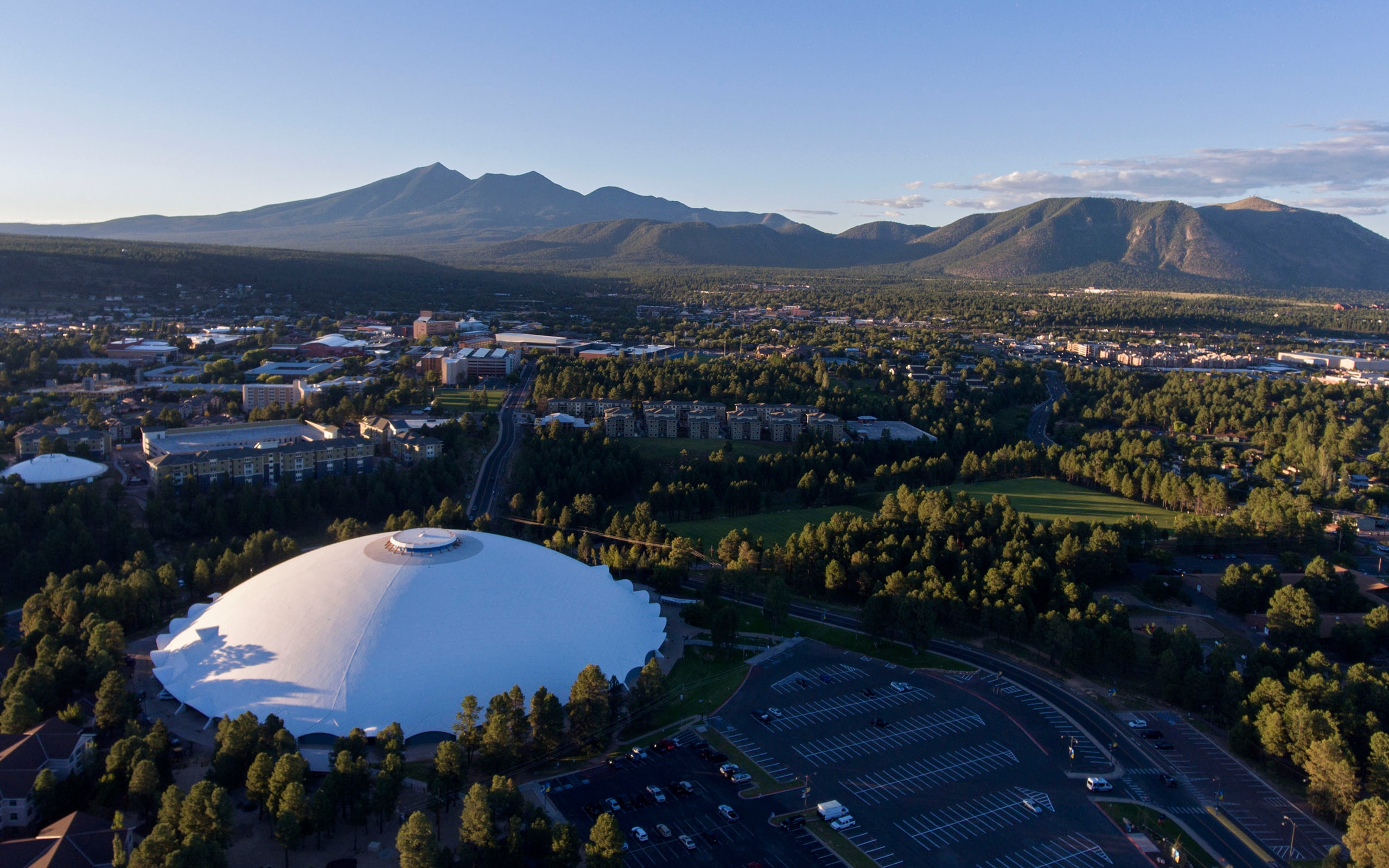Aerial view of a white domed building surrounded by trees and parking lots, with a cityscape and mountains in the background under a clear sky.