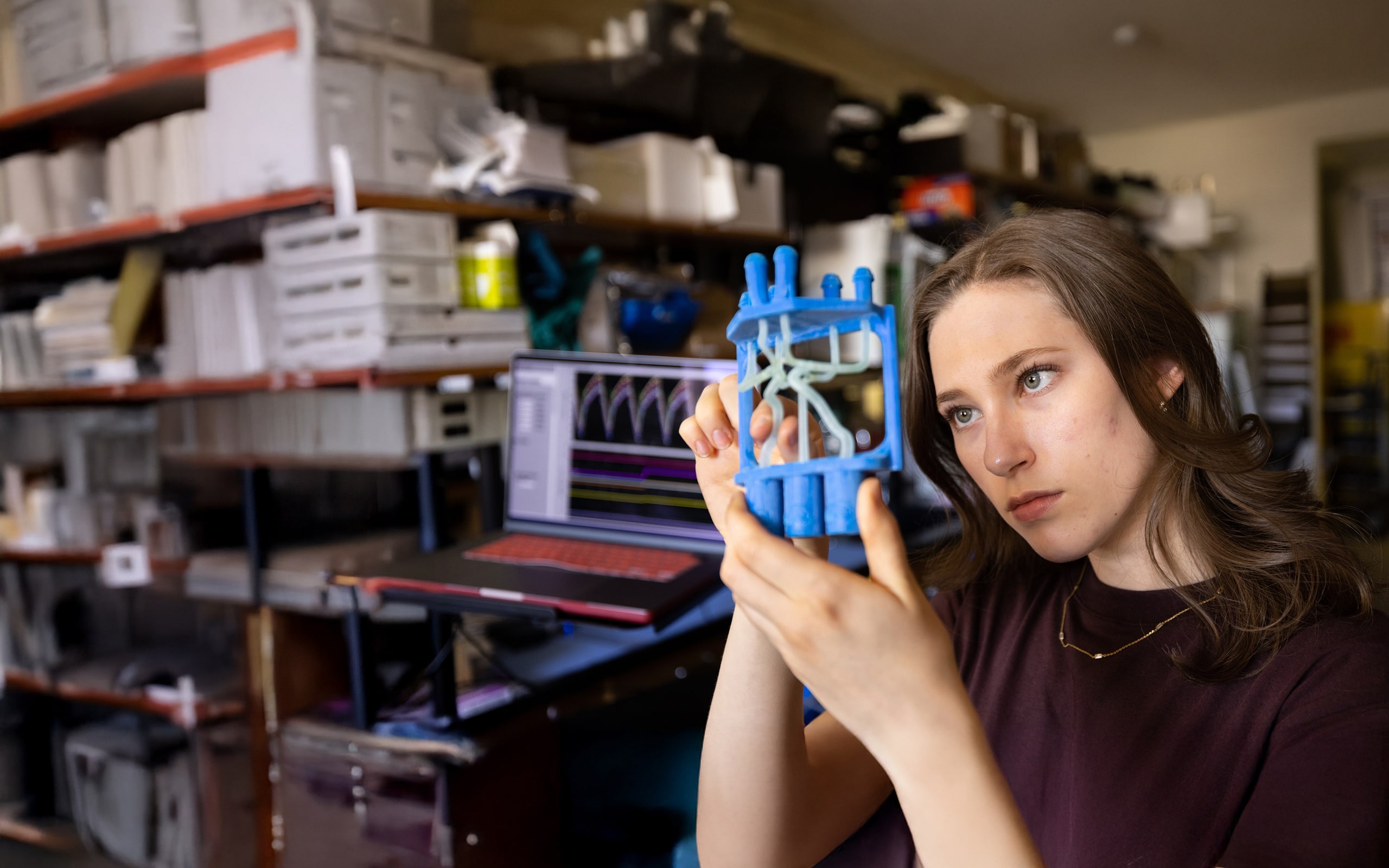A person examines a blue 3D-printed object in a workshop, with shelves of equipment and a laptop displaying a graph in the background.
