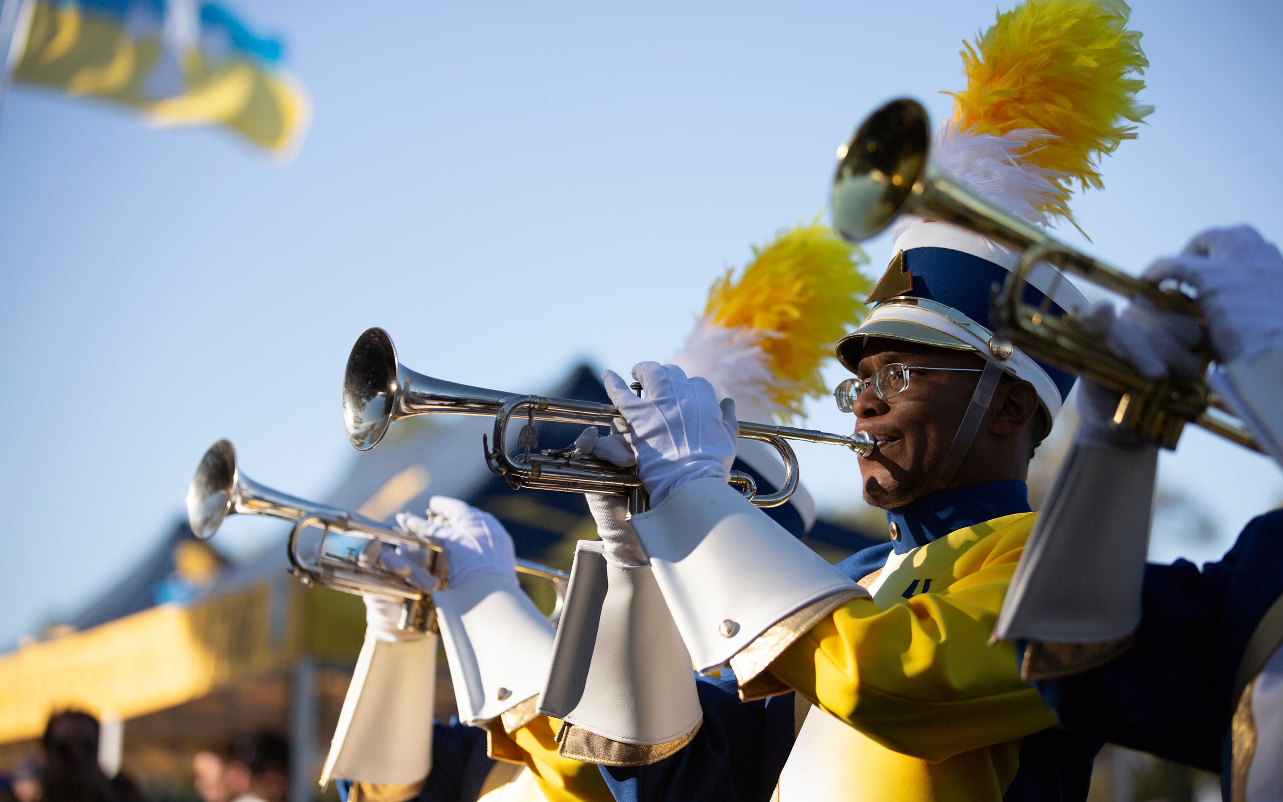 A marching band trumpet player in a blue and yellow uniform and plume hat performs outdoors with other band members.