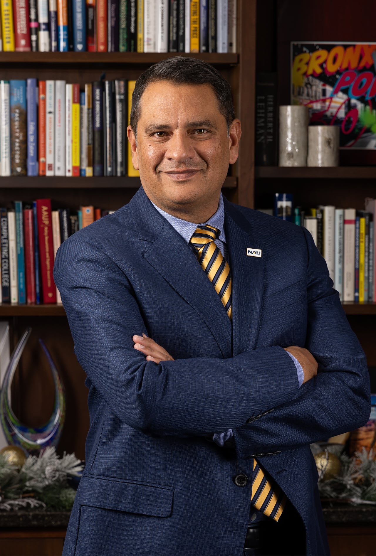 A man in a blue suit and striped tie stands with arms crossed in front of a bookshelf filled with books and decorative items.