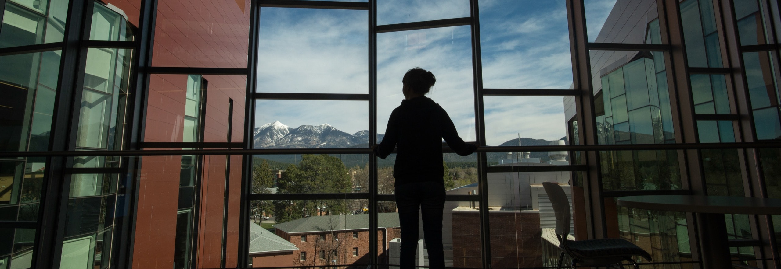 A person stands by large windows in a modern building, looking out at snow-capped mountains and a blue sky with scattered clouds.