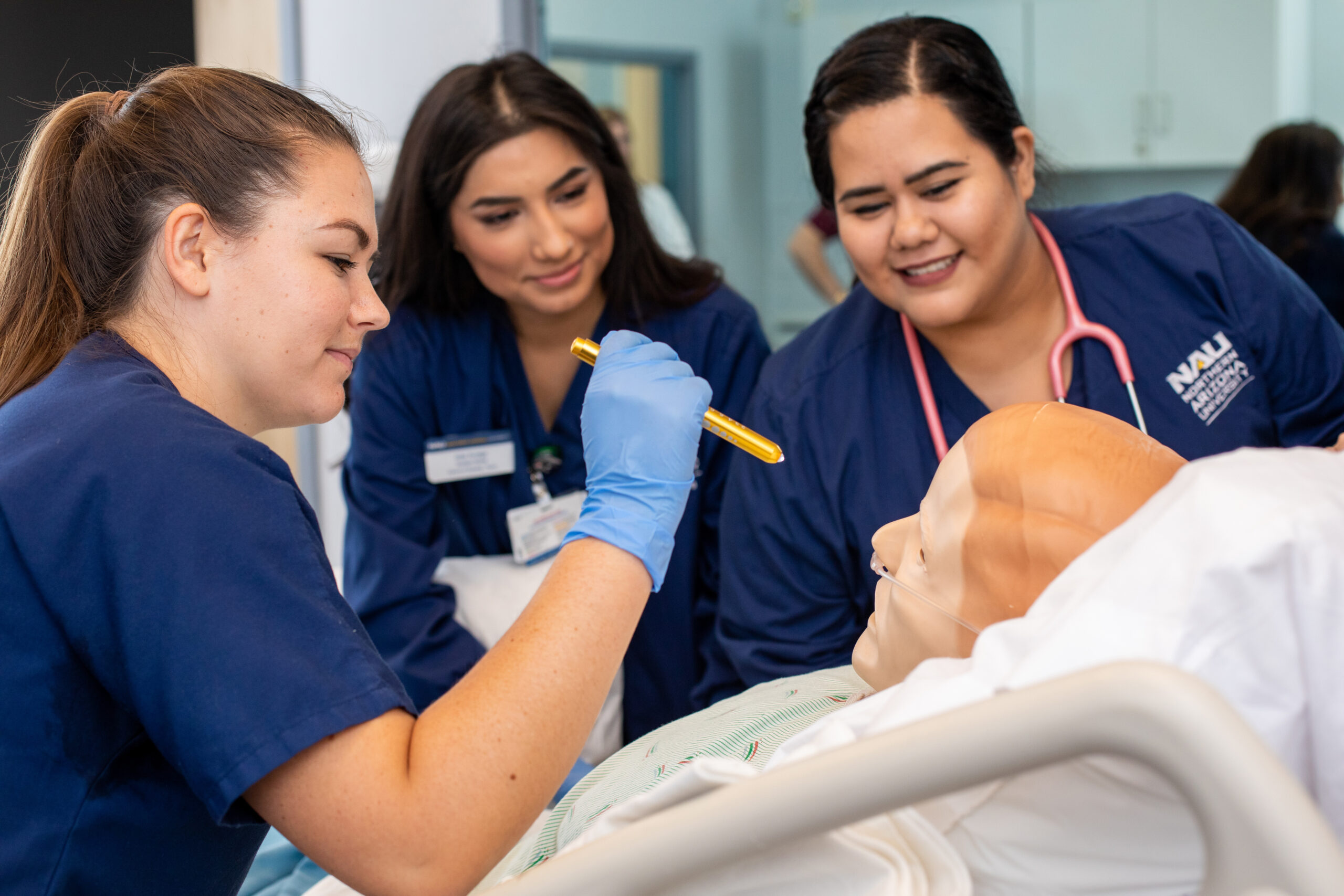 Three nursing students in blue uniforms practice a medical procedure on a simulation mannequin in a clinical training setting.