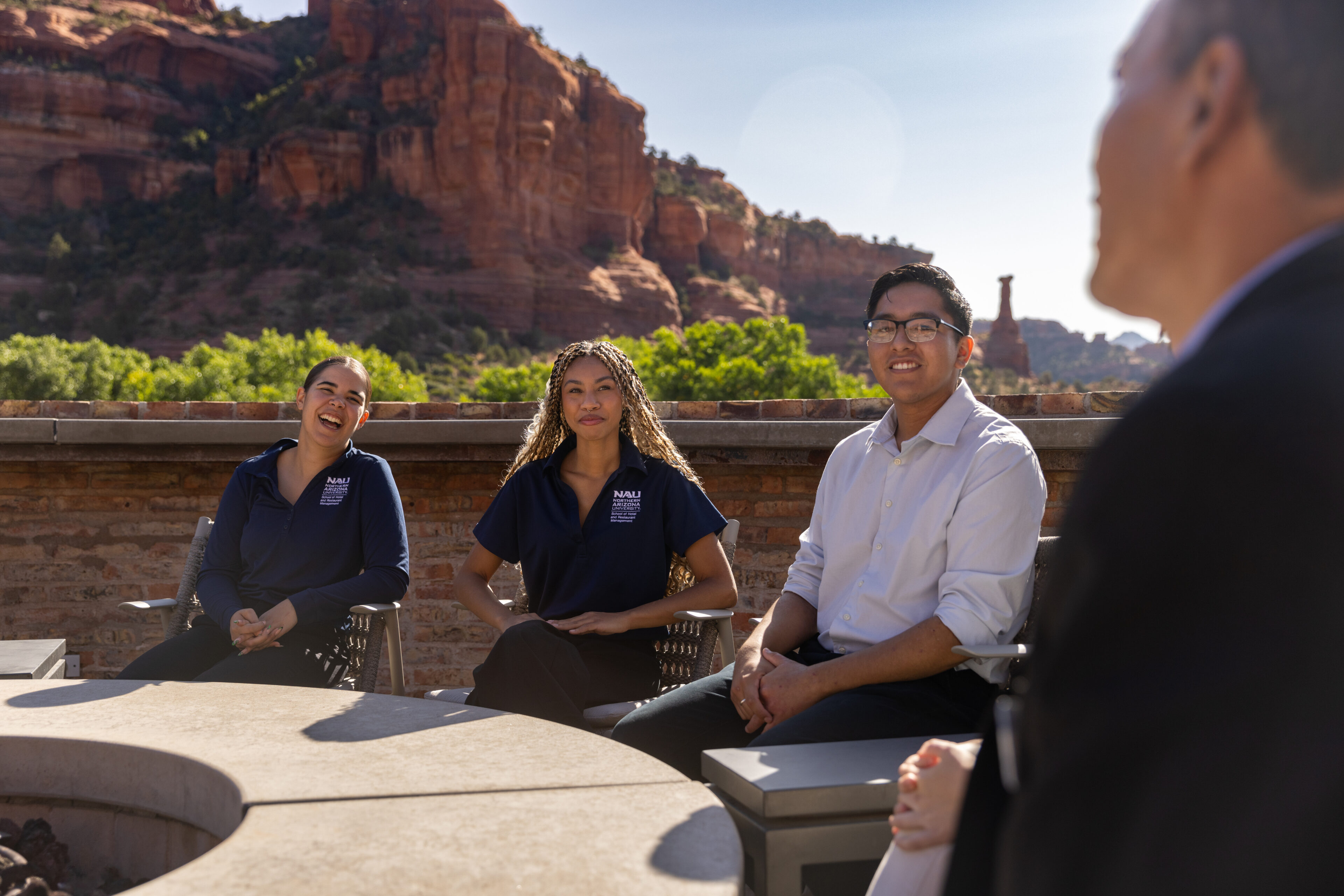 Four people sit around an outdoor table with red rock cliffs in the background, engaged in conversation on a sunny day.