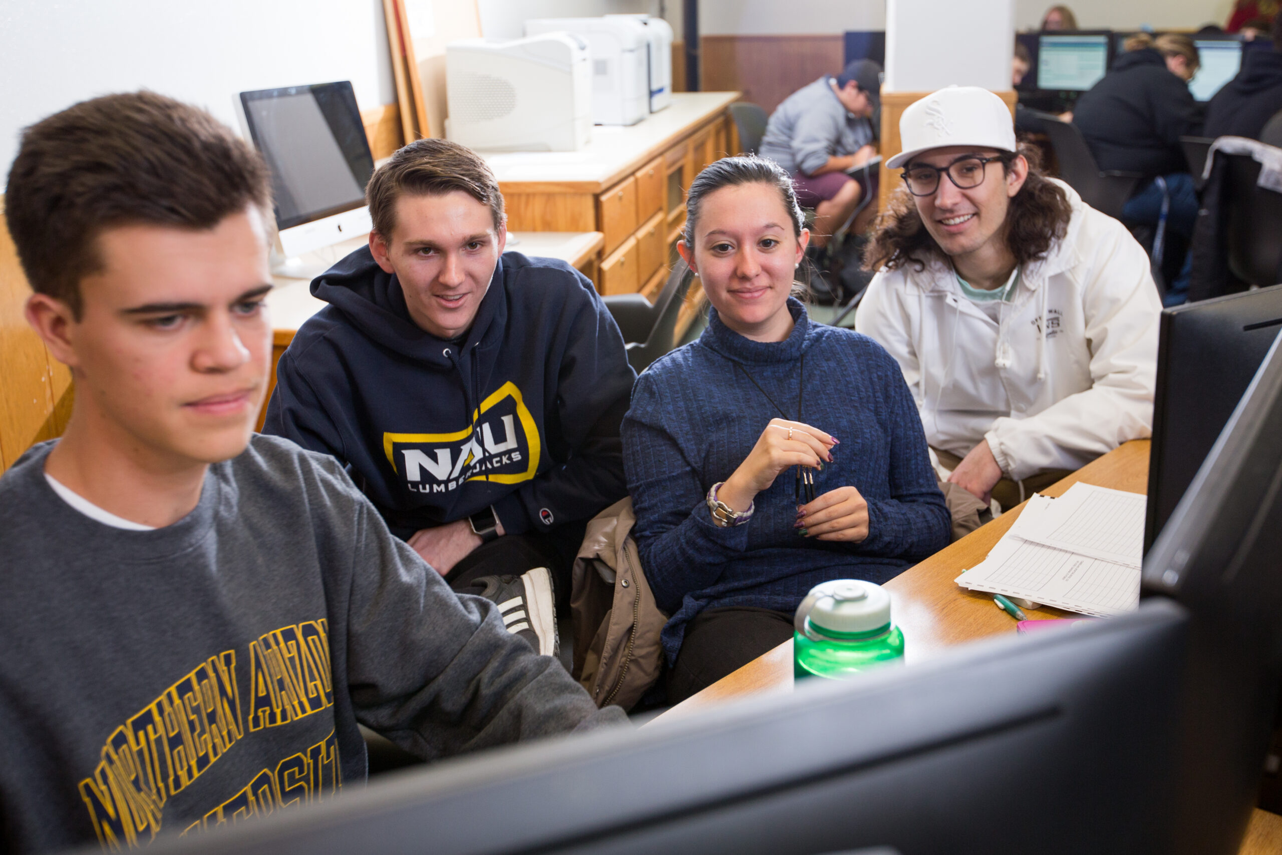 Four college students sit together at a desk, working on a computer in a classroom setting with papers and a water bottle nearby.
