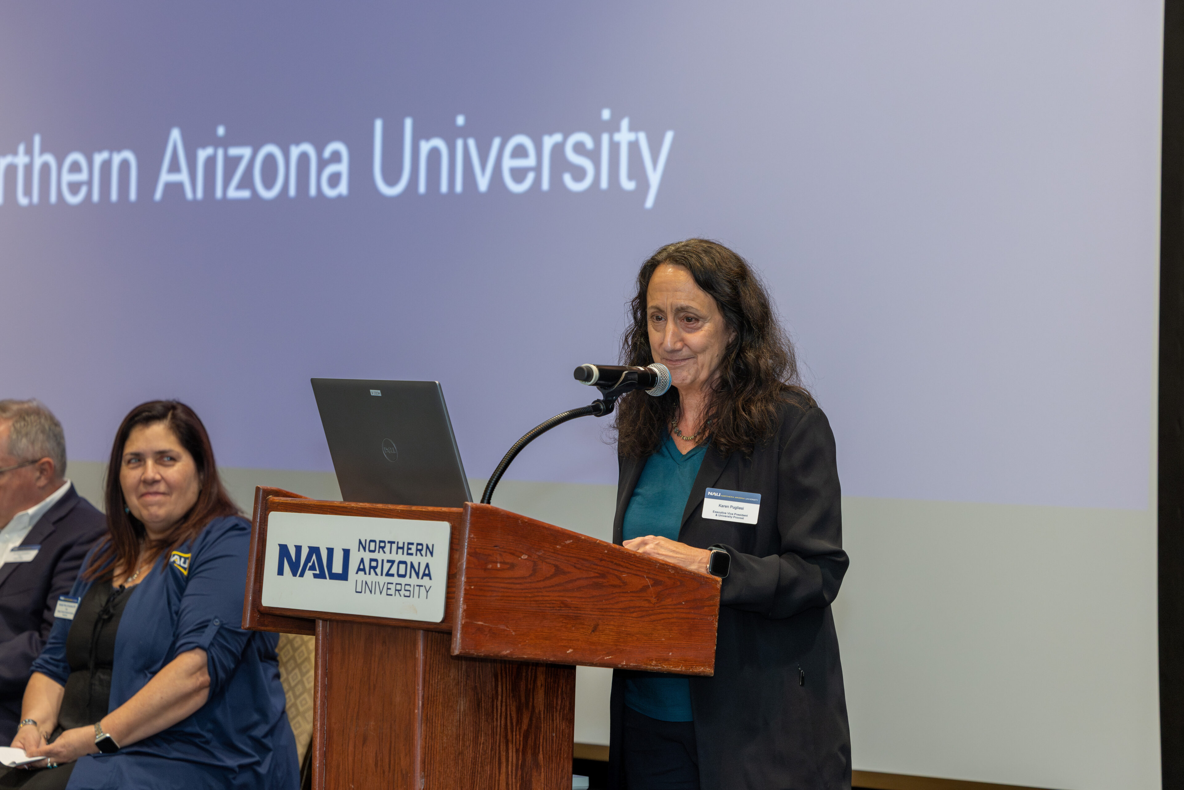 A woman stands at a podium labeled "Northern Arizona University," speaking into a microphone while others sit nearby on a panel.