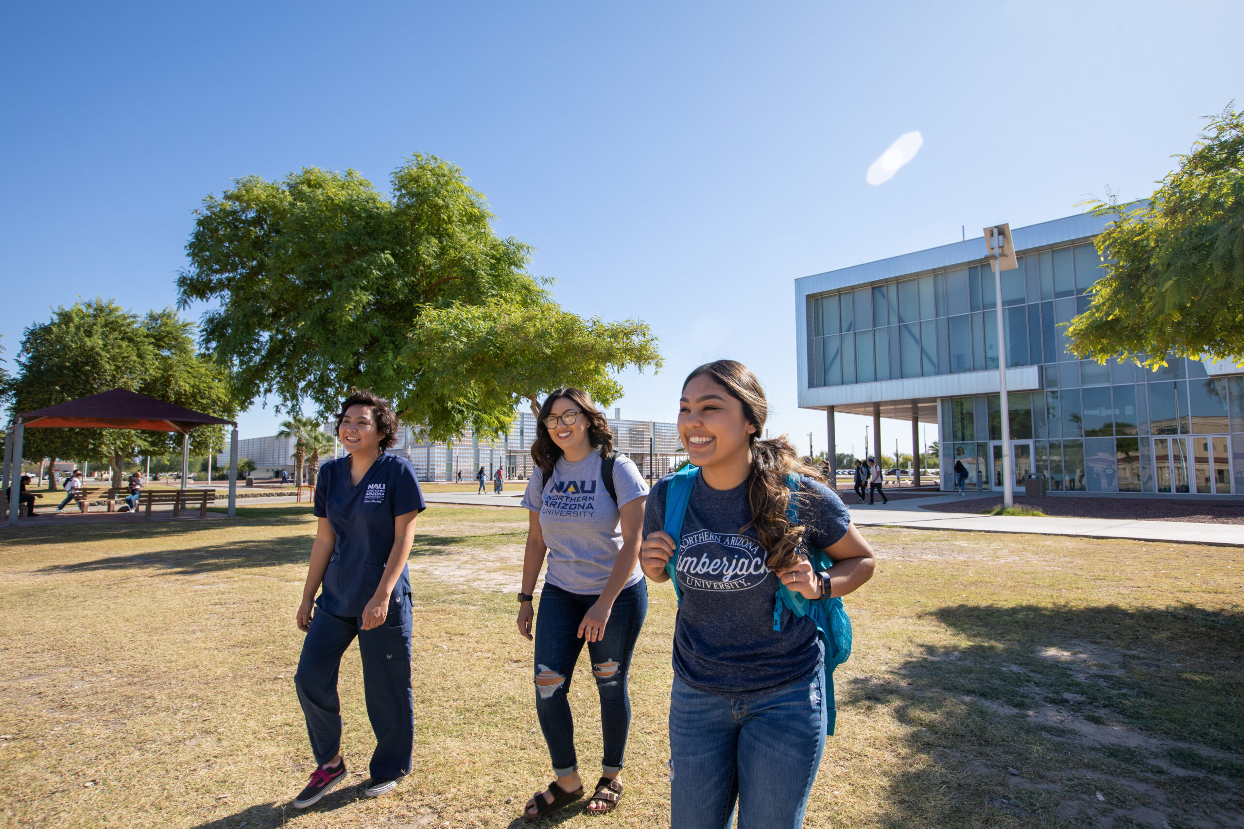 Three students walk together outside on a sunny day near a modern campus building, with trees and grass surrounding them.