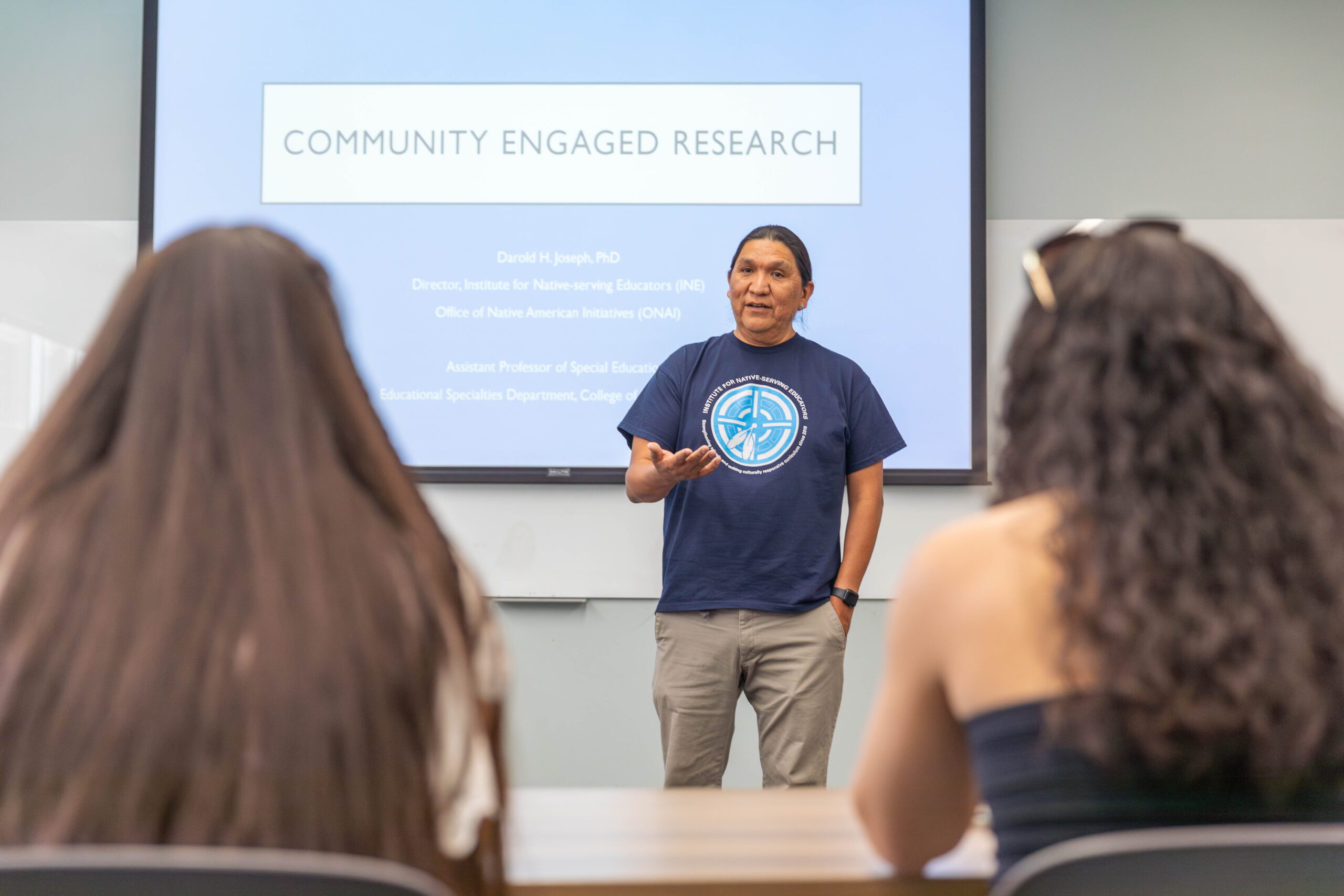 A man stands in front of a presentation screen titled "Community Engaged Research," speaking to two seated attendees in a classroom setting.