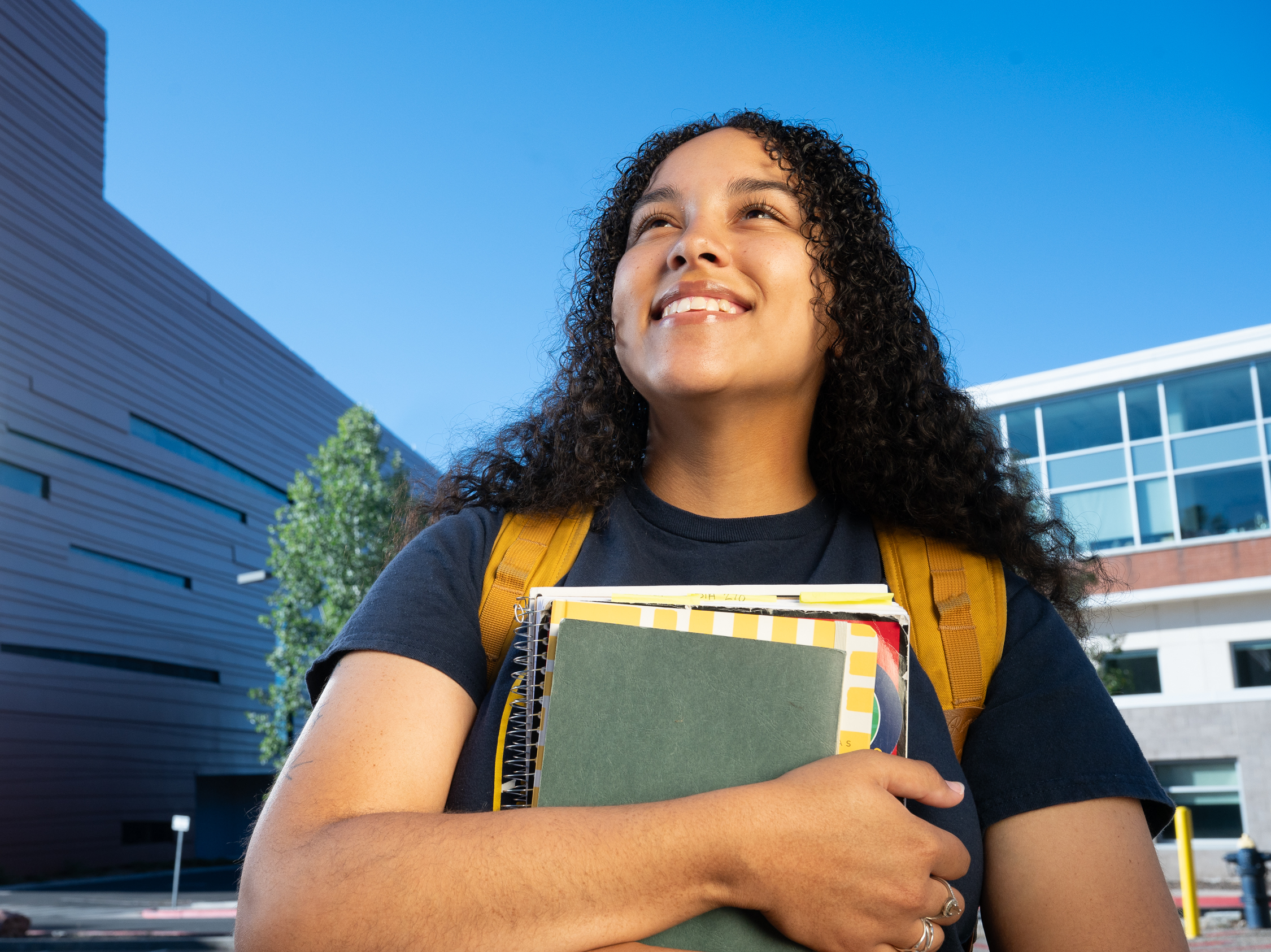Young woman with curly hair holds notebooks and smiles outside a modern school building on a sunny day.