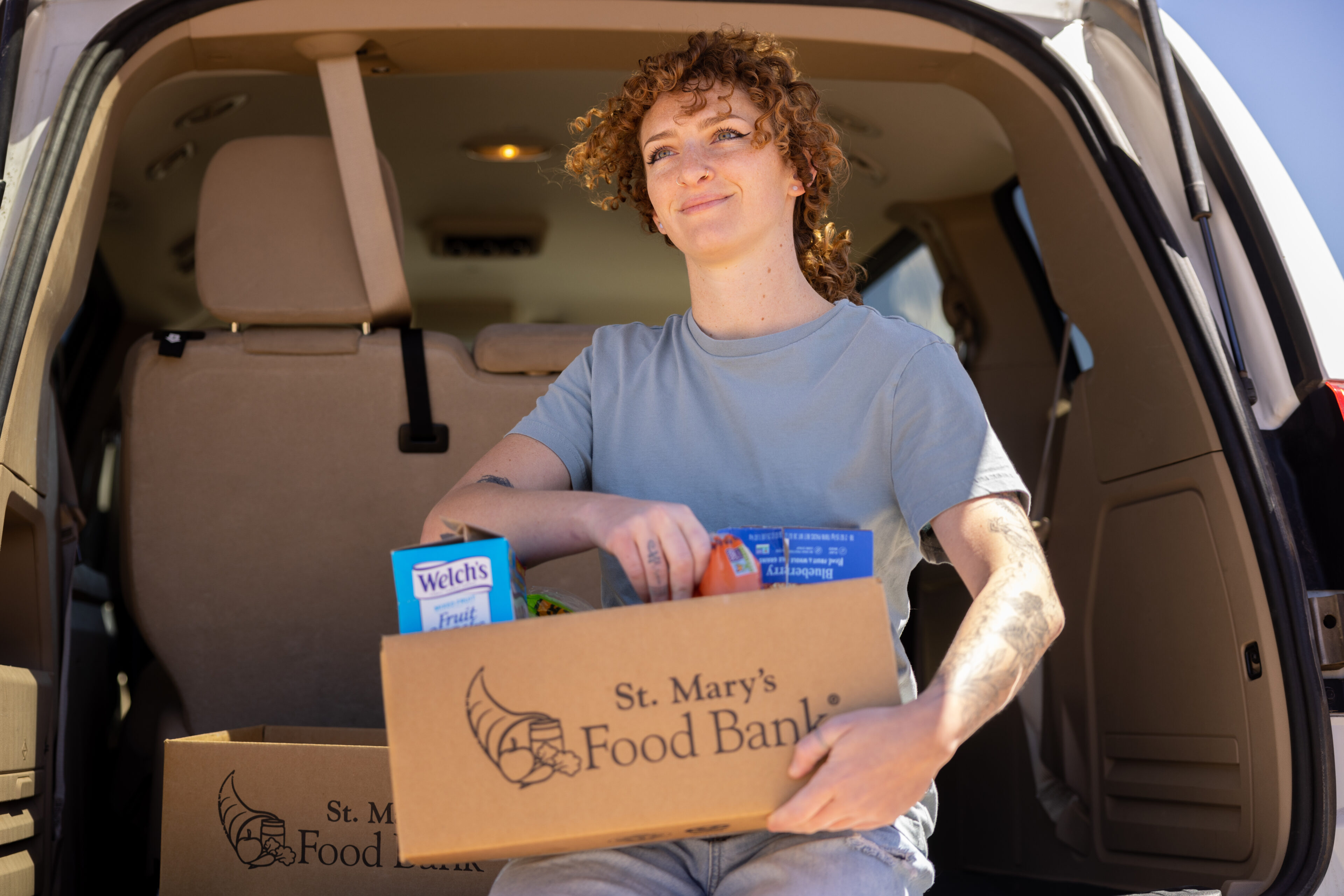 A person sits in the back of a vehicle holding a St. Mary's Food Bank box filled with groceries, including fruit snacks and canned goods.