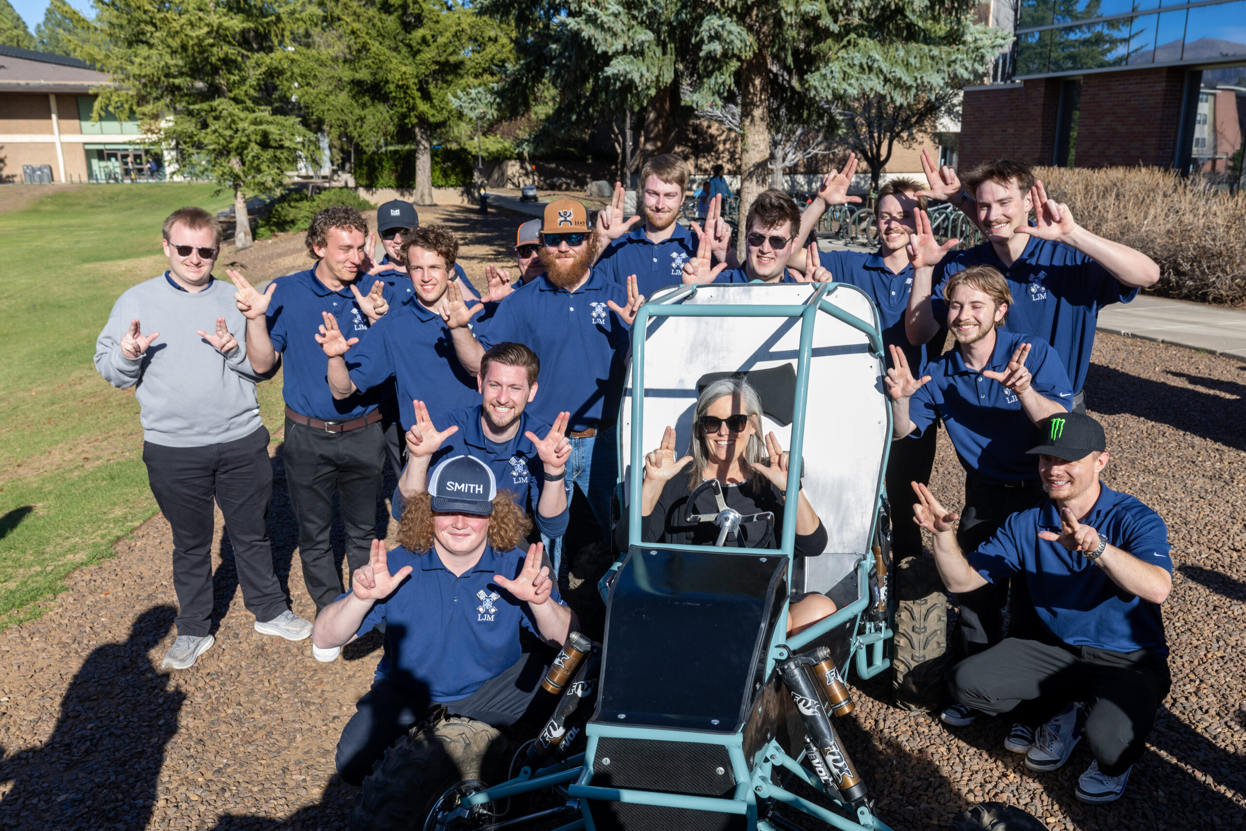 A group of people in matching blue shirts pose outdoors around a small open-frame vehicle, many holding up "L" hand signs.