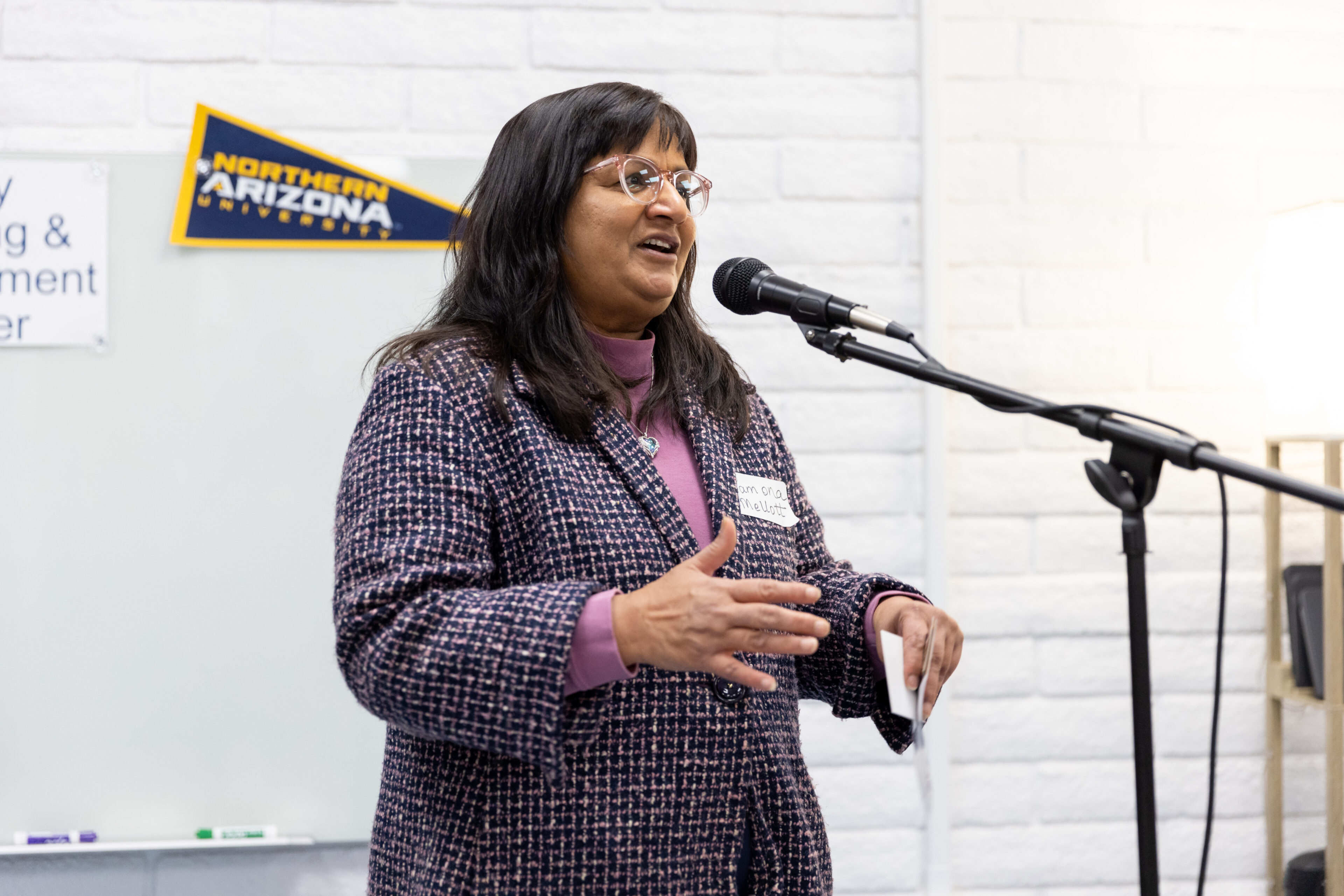 A woman speaks into a microphone while standing in front of a whiteboard, with a Northern Arizona University pennant visible on the wall.