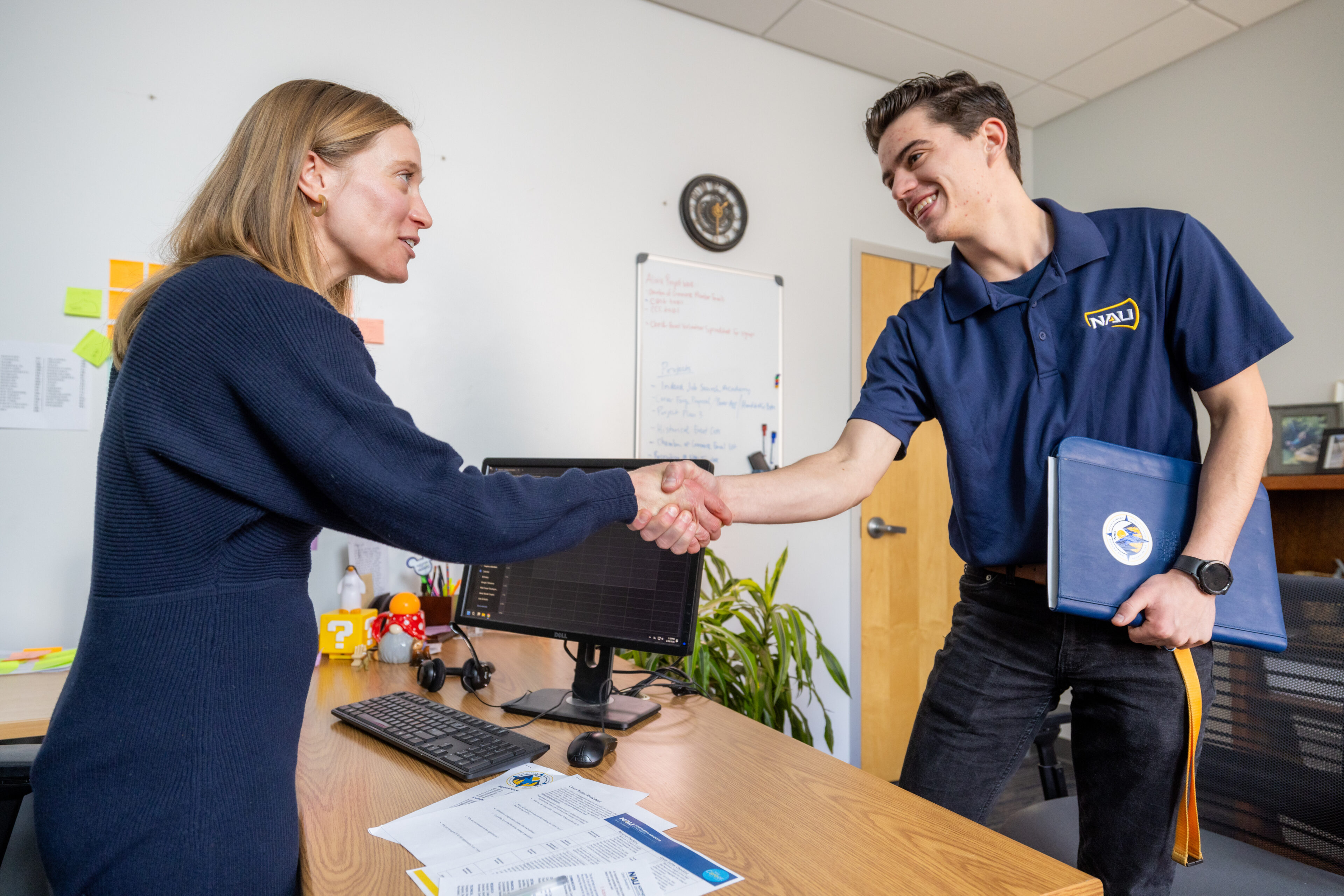 A woman and a man shake hands in an office. The man holds a folder and wears a polo shirt with a logo. They are standing next to a desk with office supplies and paperwork.
