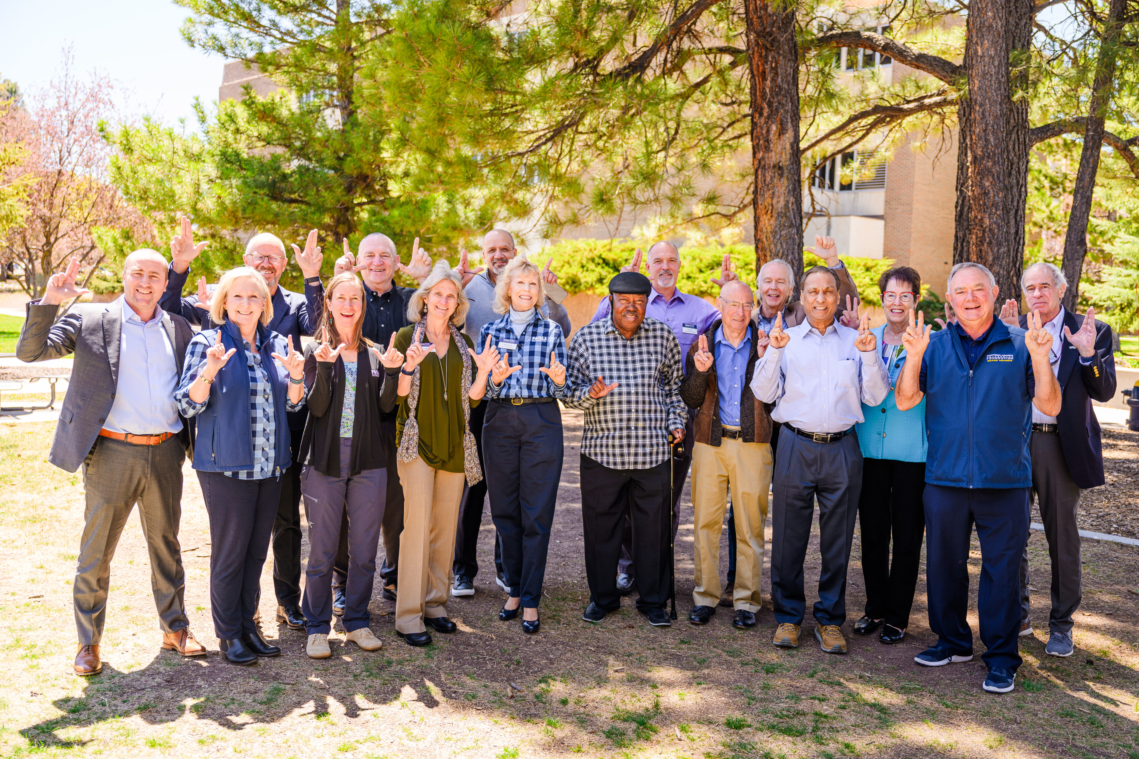 Group of adults standing outdoors on grass, posing together and making hand gestures, with trees and a building in the background.