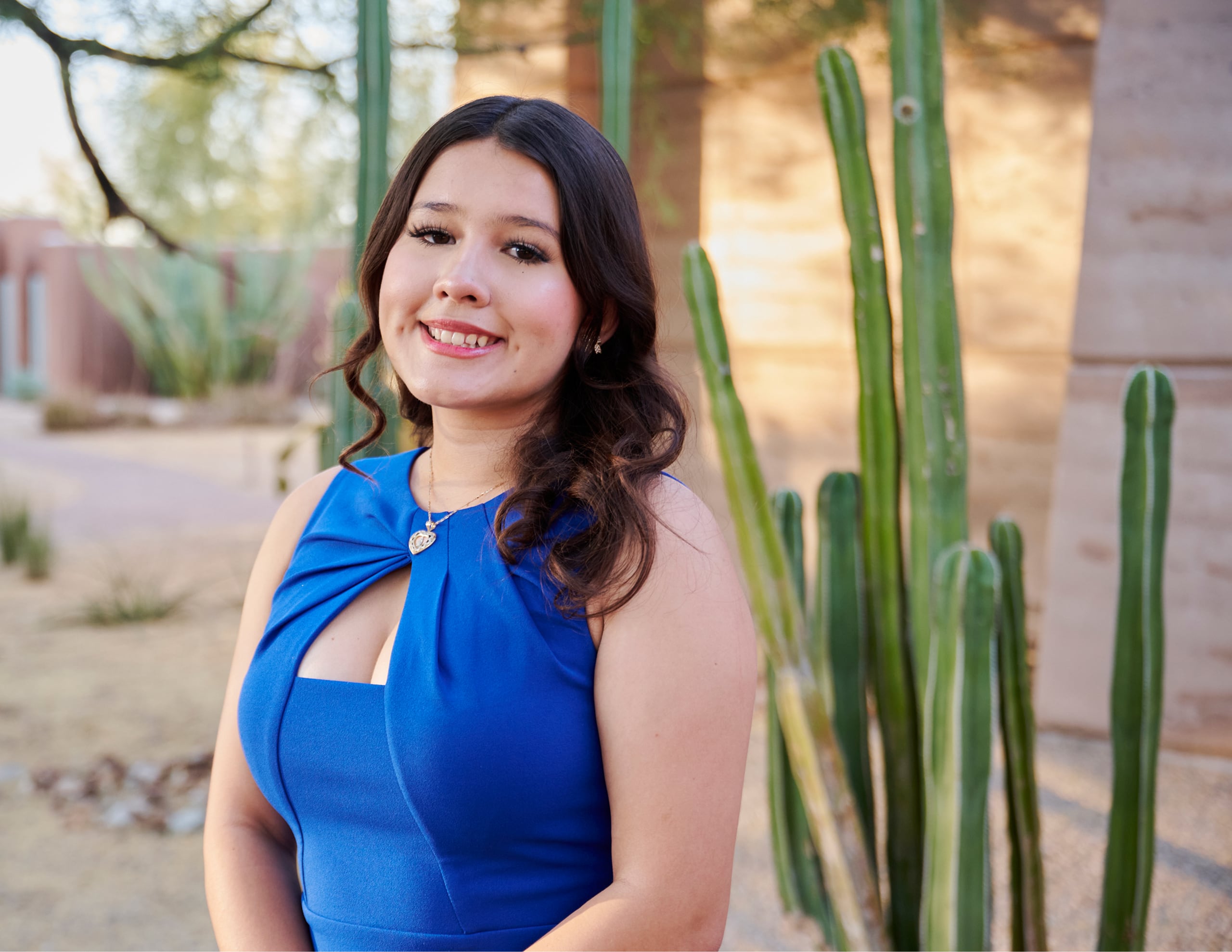 A young woman in a blue dress stands outdoors in front of tall cacti and desert landscaping, smiling at the camera.