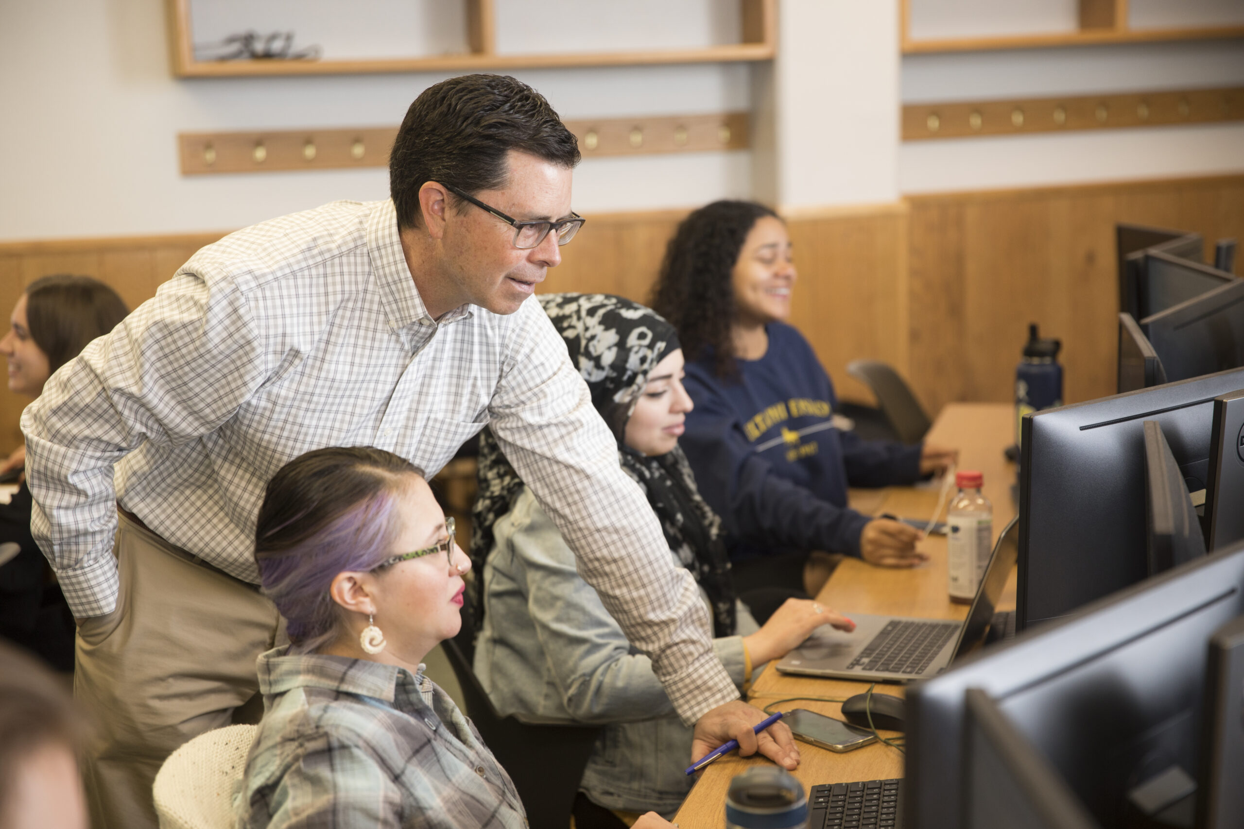 A man stands next to a woman in a hijab, assisting her at a computer, while other students work at their computers in a classroom.