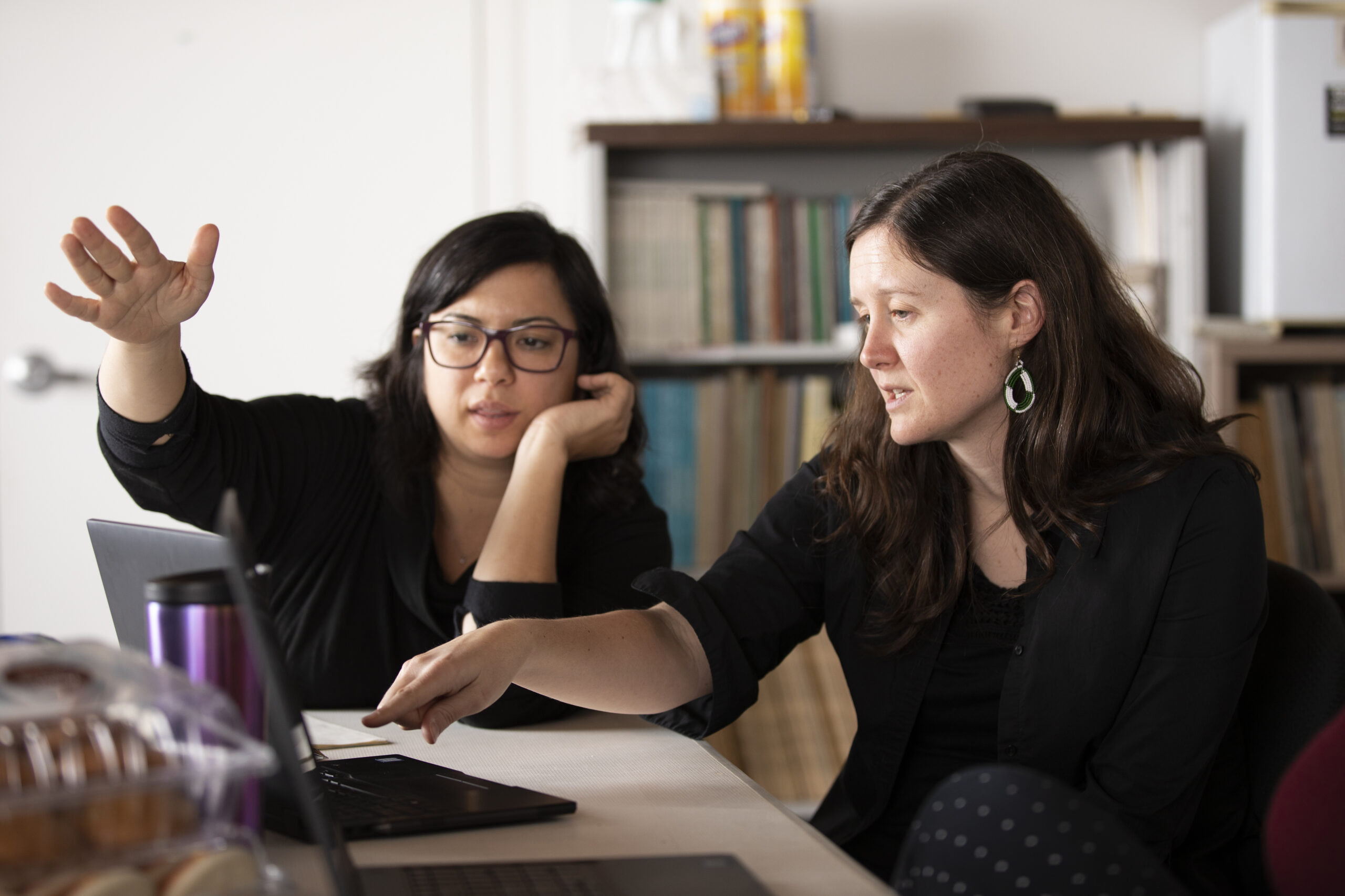 Two women sitting at a table, engaged in discussion while looking at a laptop; one gestures with her hand while the other points at the screen.
