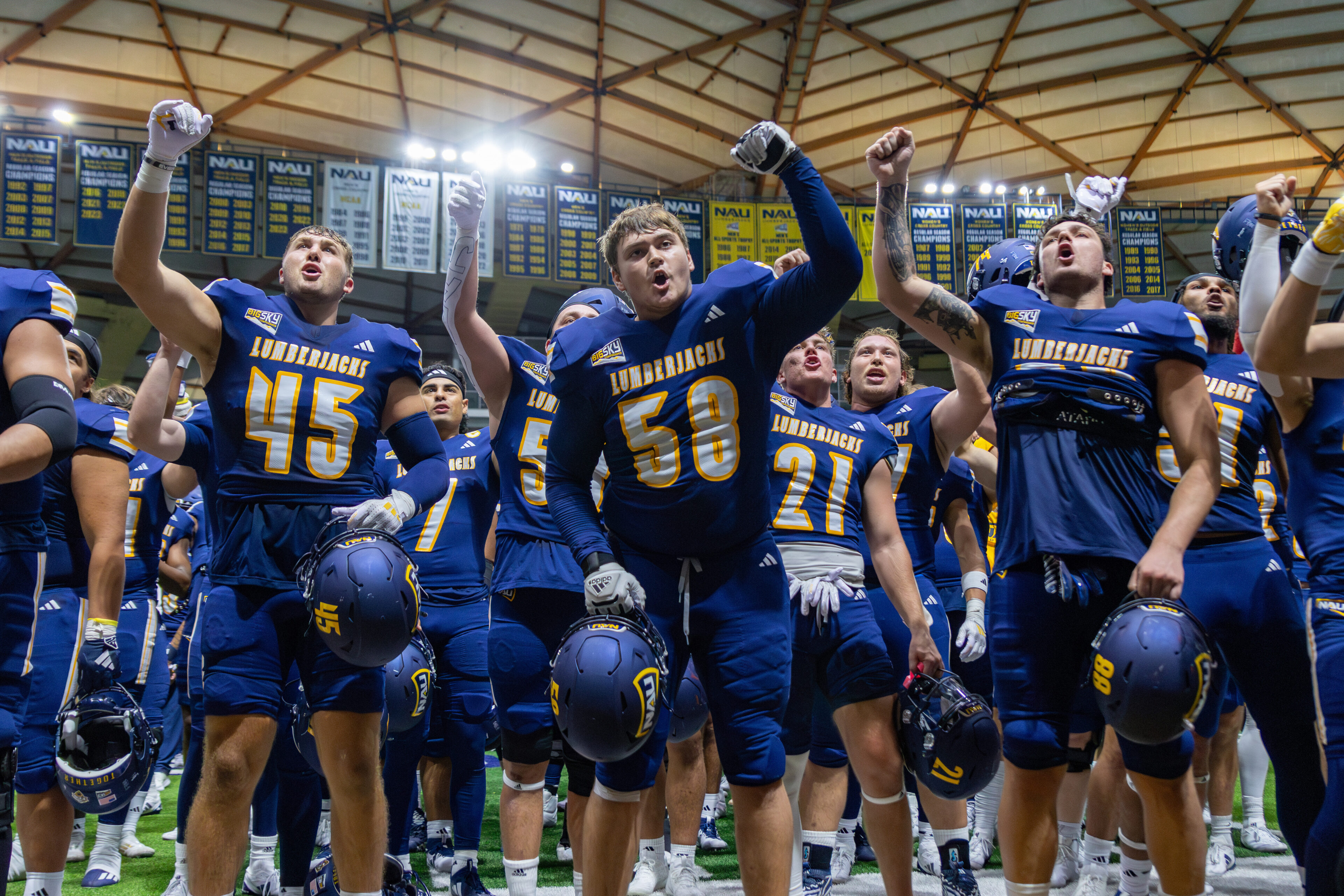 Football players in blue and yellow jerseys, helmets, and uniforms celebrating together on a sports field with stadium seating visible in the background.