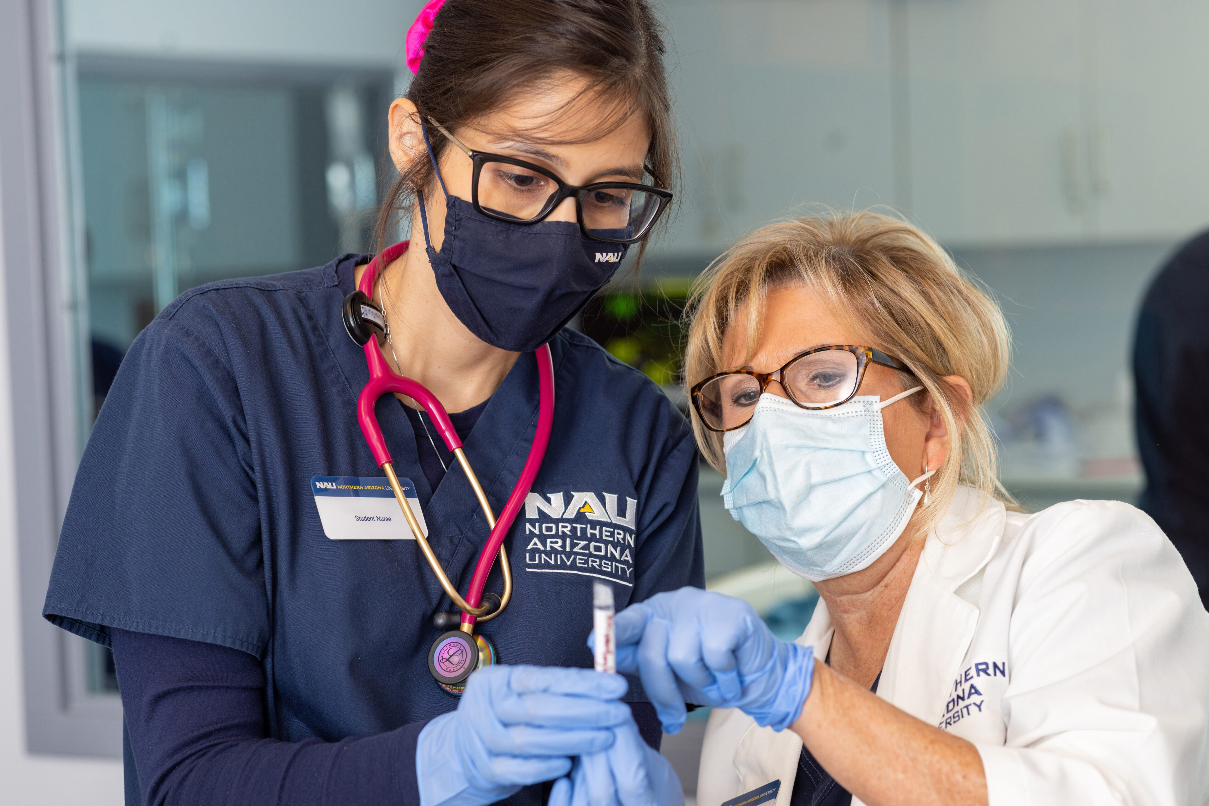 Two healthcare professionals in masks and gloves prepare a syringe in a clinical setting. One wears NAU scrubs, while the other wears a white lab coat.