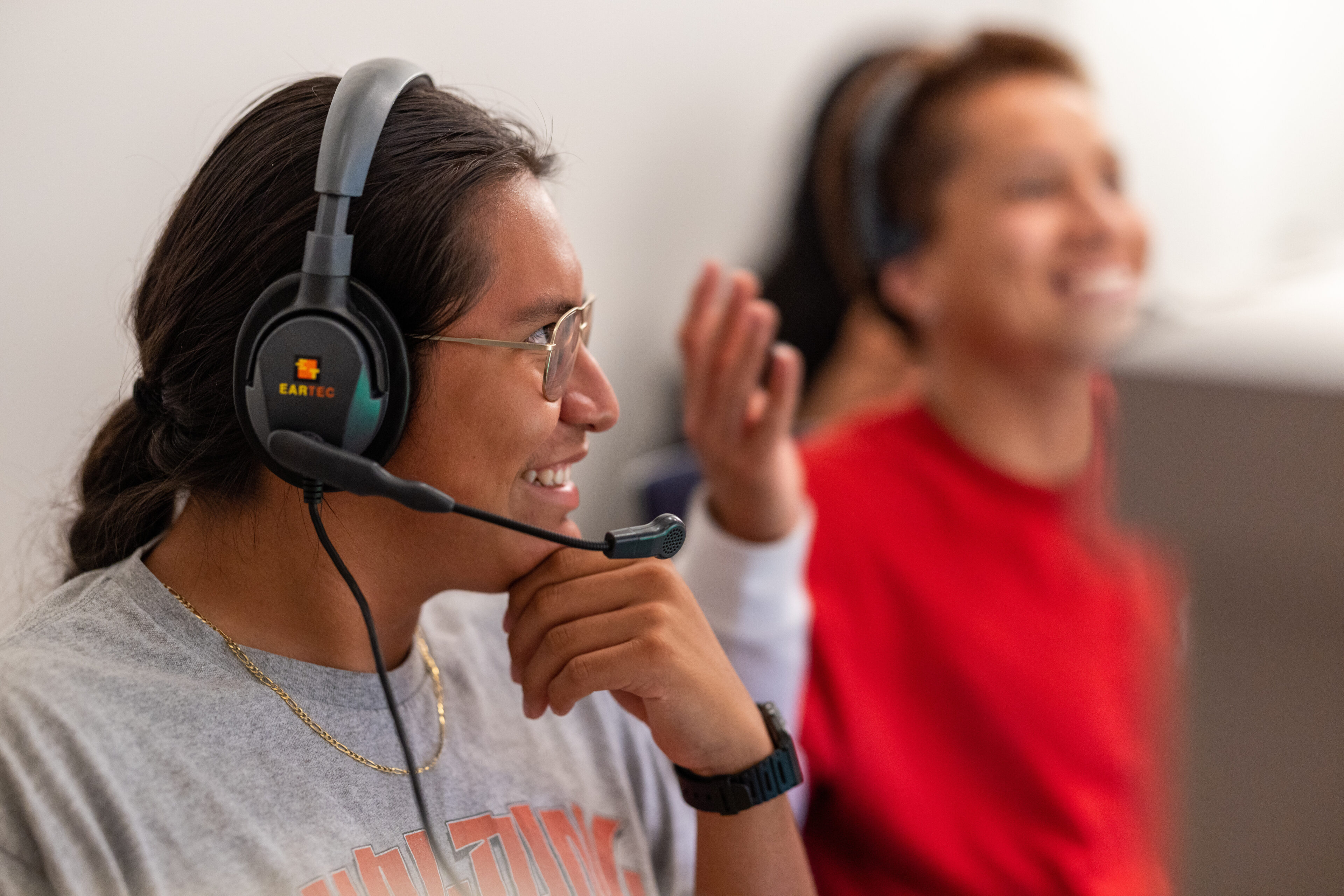 Two people wearing headsets sit indoors; one in focus, smiling and speaking, while the other is blurred in the background, gesturing with their hand.