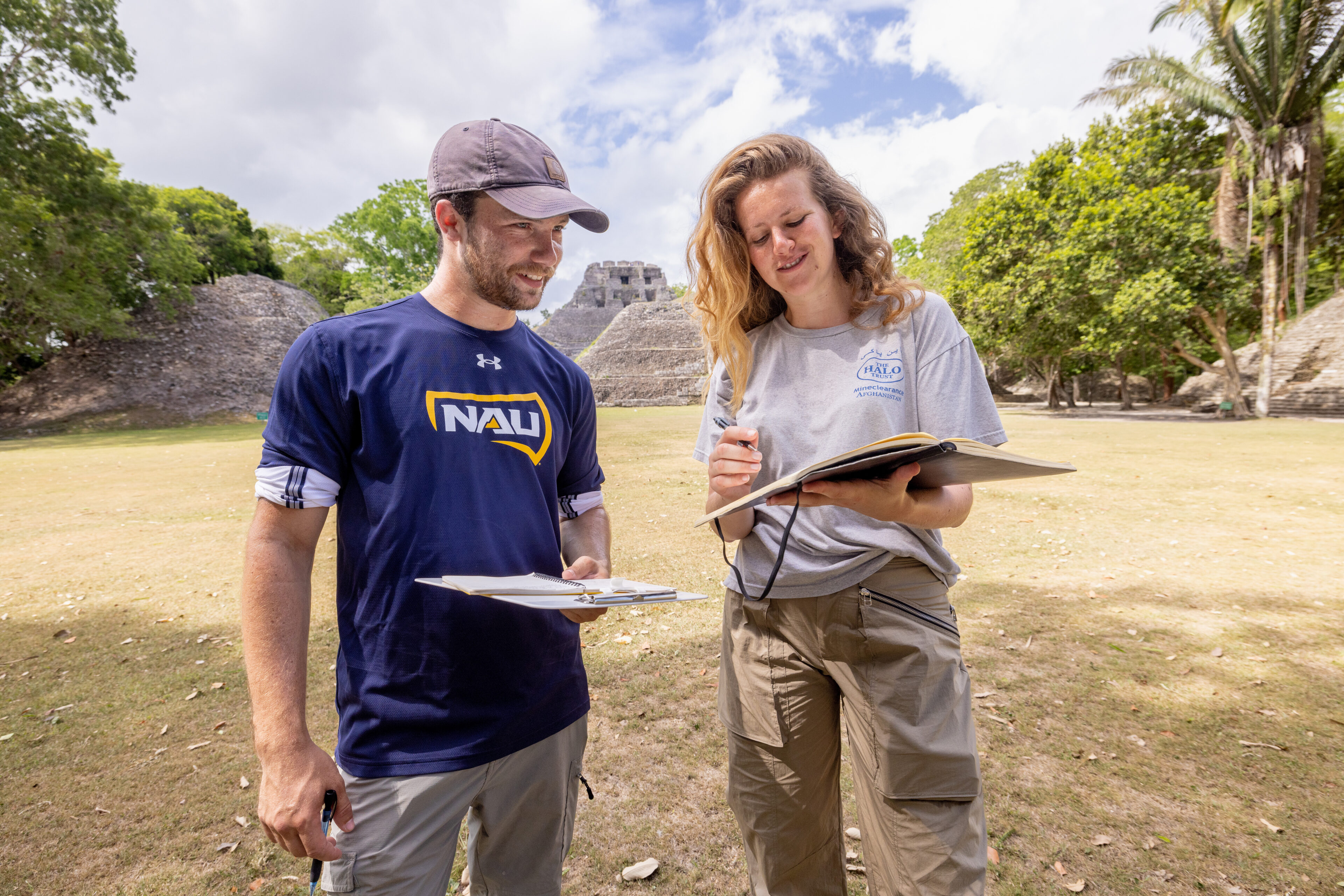 Two people stand outdoors with clipboards and notebooks, recording information near ancient stone ruins and trees under a partly cloudy sky.