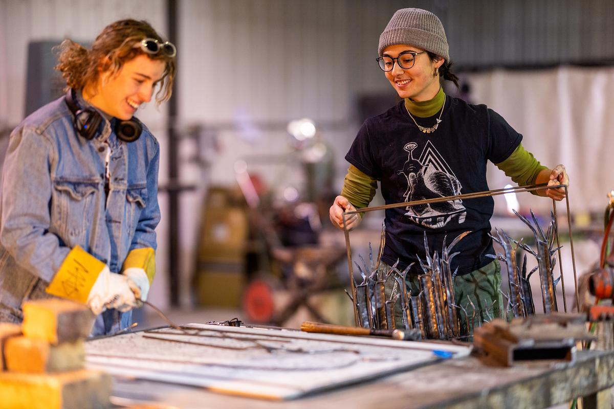 Two people in casual clothes work together on a metal art project in a workshop, handling metal rods and tools on a large table.