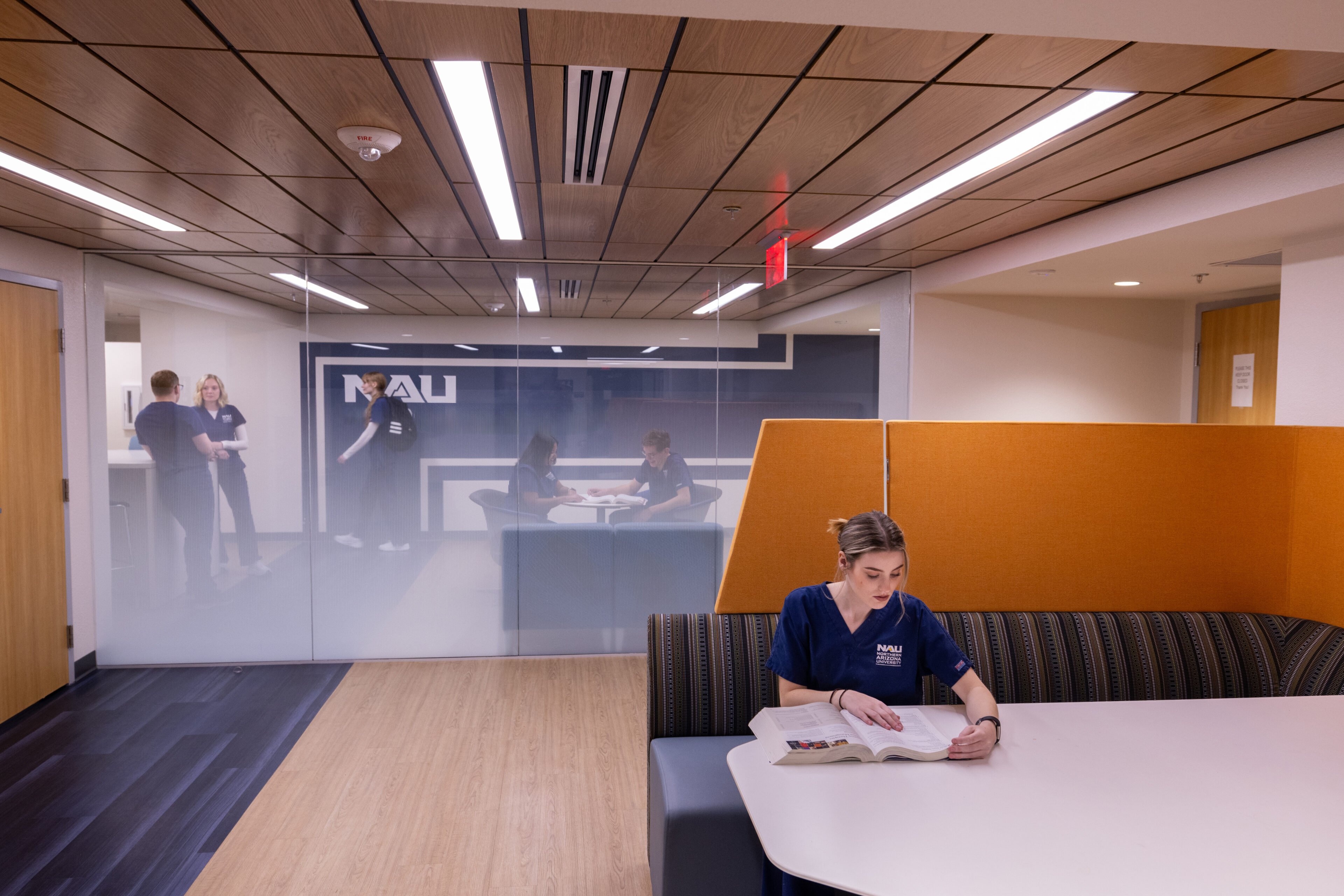 A woman in scrubs reads a textbook at a table in a modern study area while two others stand and talk near a glass wall in the background.