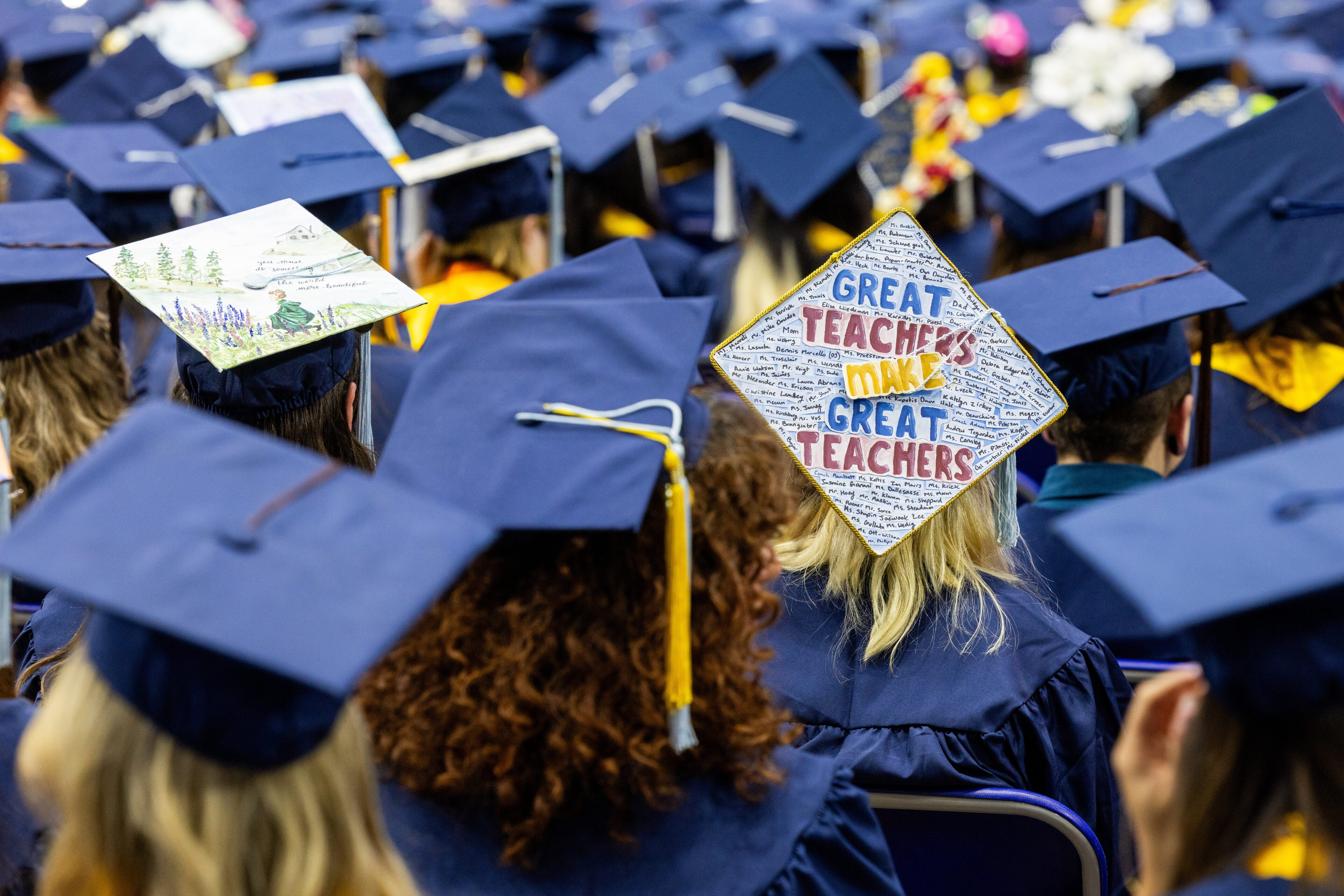 Graduates in navy caps and gowns are seated at a ceremony; one cap is decorated with the phrase "Great teachers make great teachers.