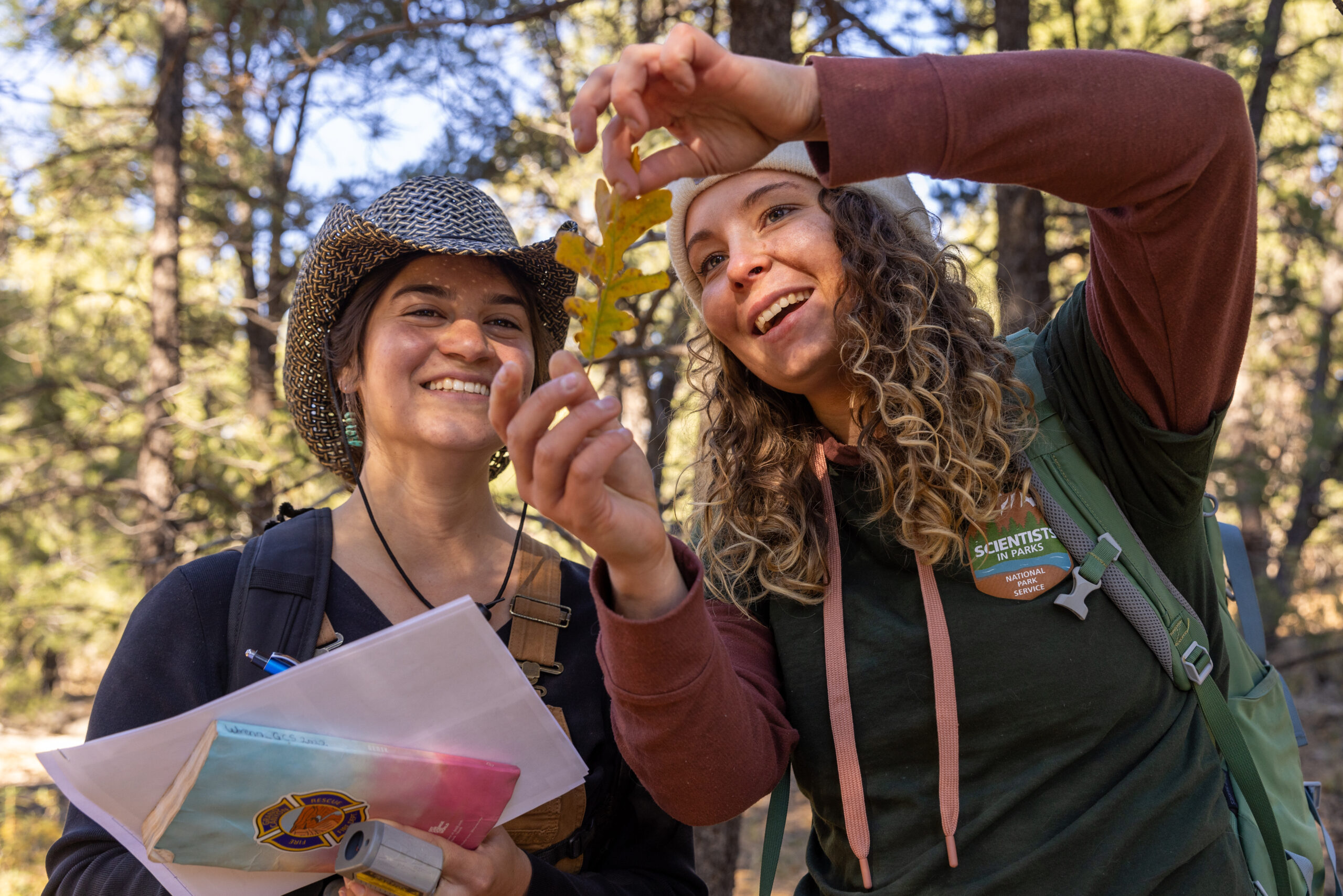 Two women outdoors examine a leaf; one holds papers and a pen, while the other holds up the leaf. Both are smiling and surrounded by trees.