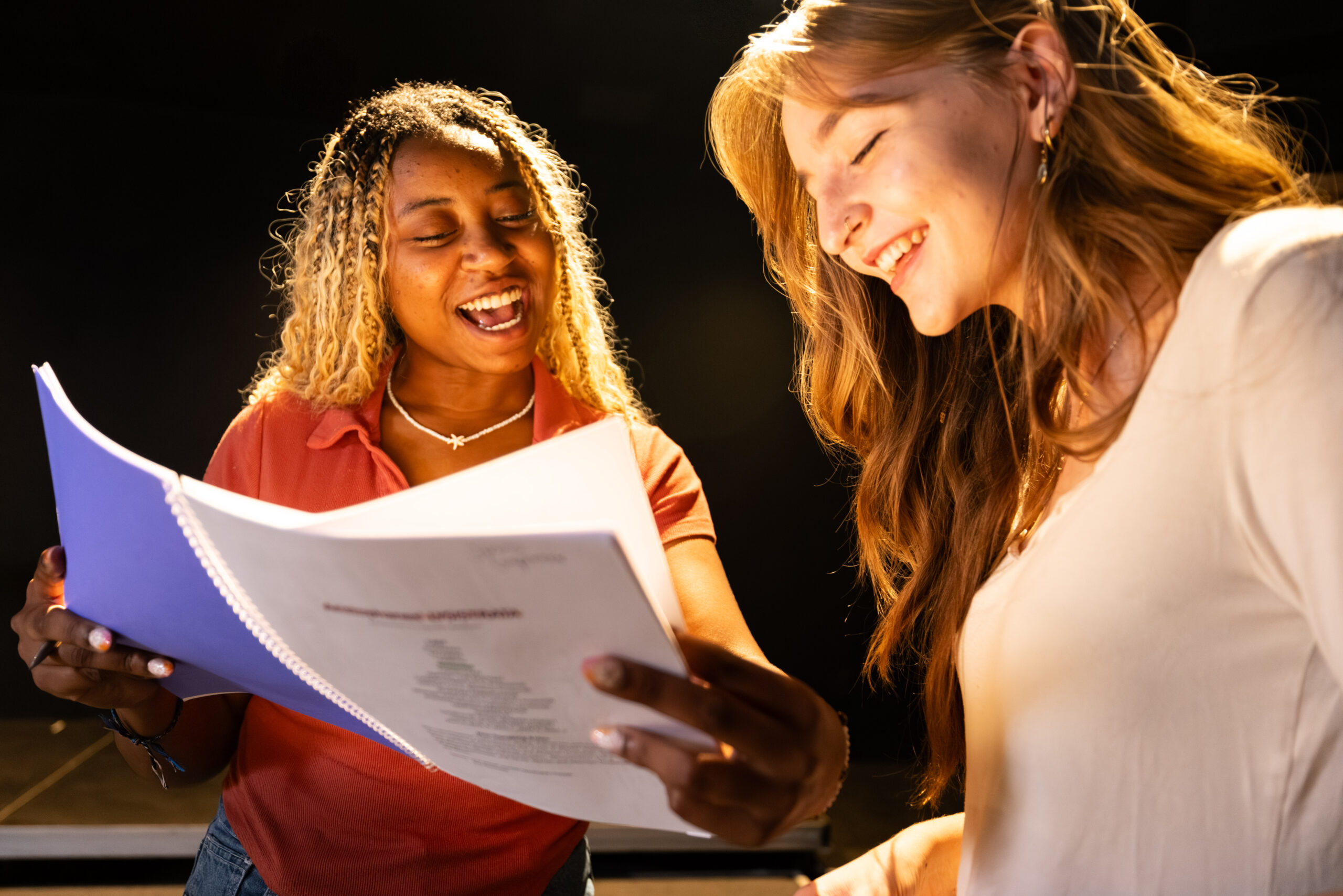 Two women stand together under bright lighting, smiling as they look at and discuss the contents of a spiral-bound script or document.