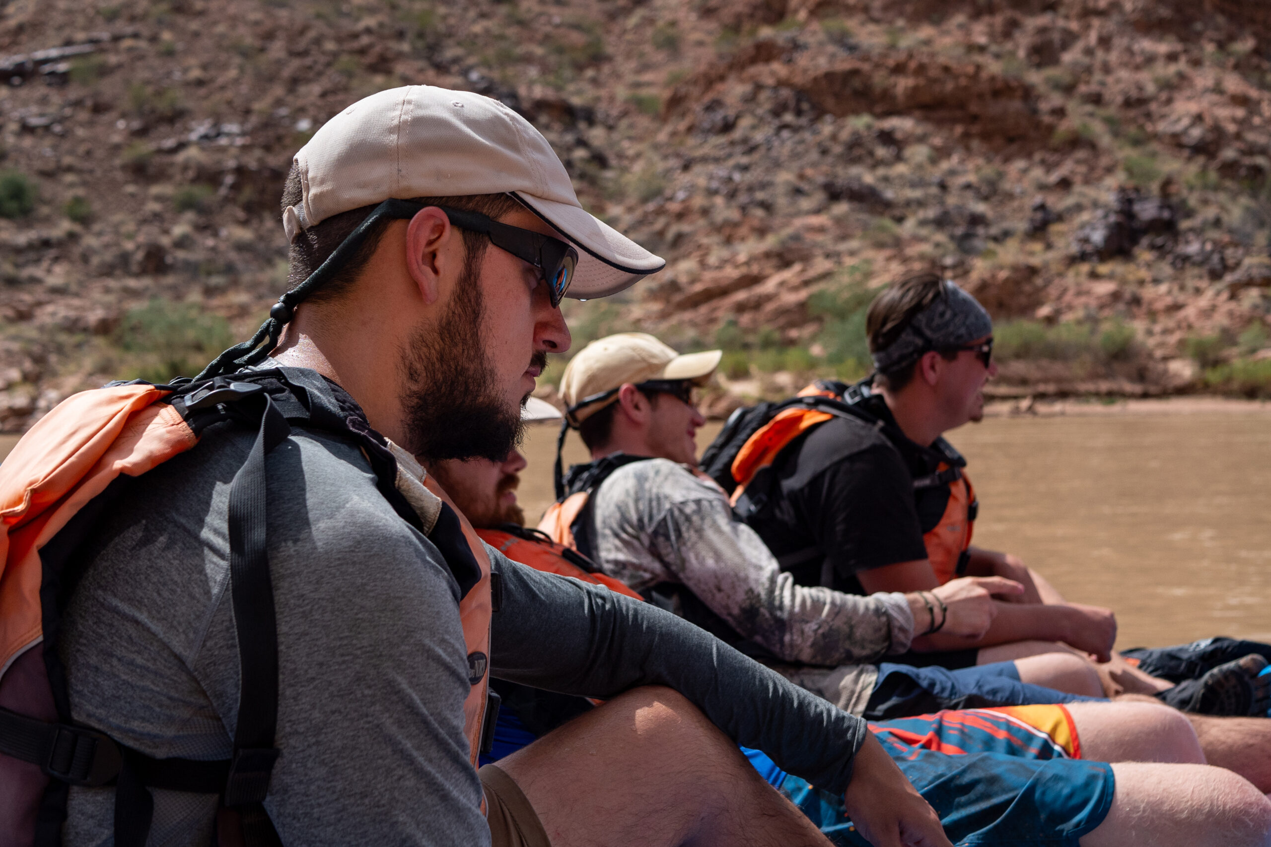 Four people wearing life jackets and caps sit in a row on a boat, with rocky terrain and muddy water visible in the background.