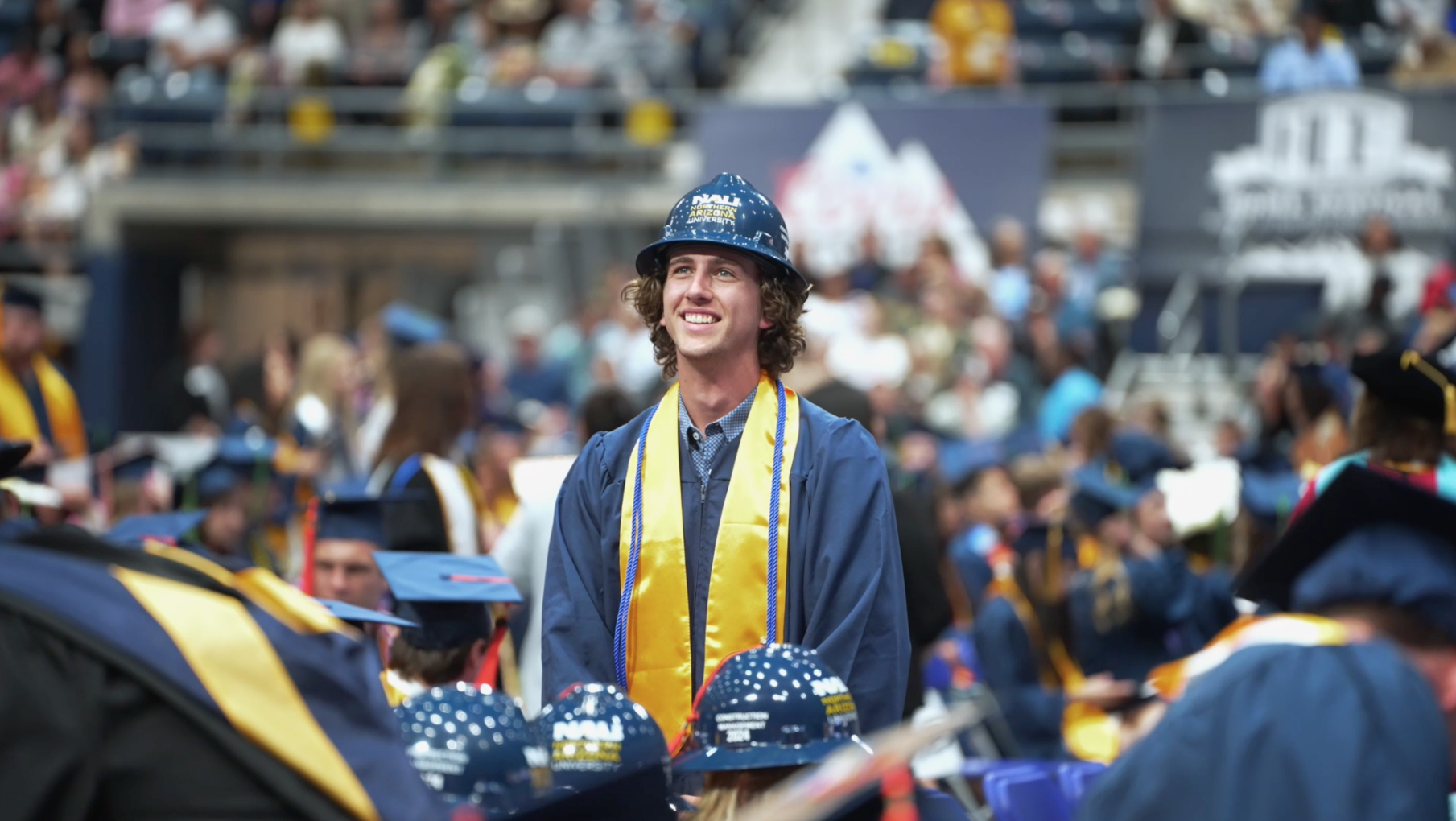 A graduate wearing a blue gown, yellow stole, and blue hard hat stands and smiles among other seated graduates at a commencement ceremony.
