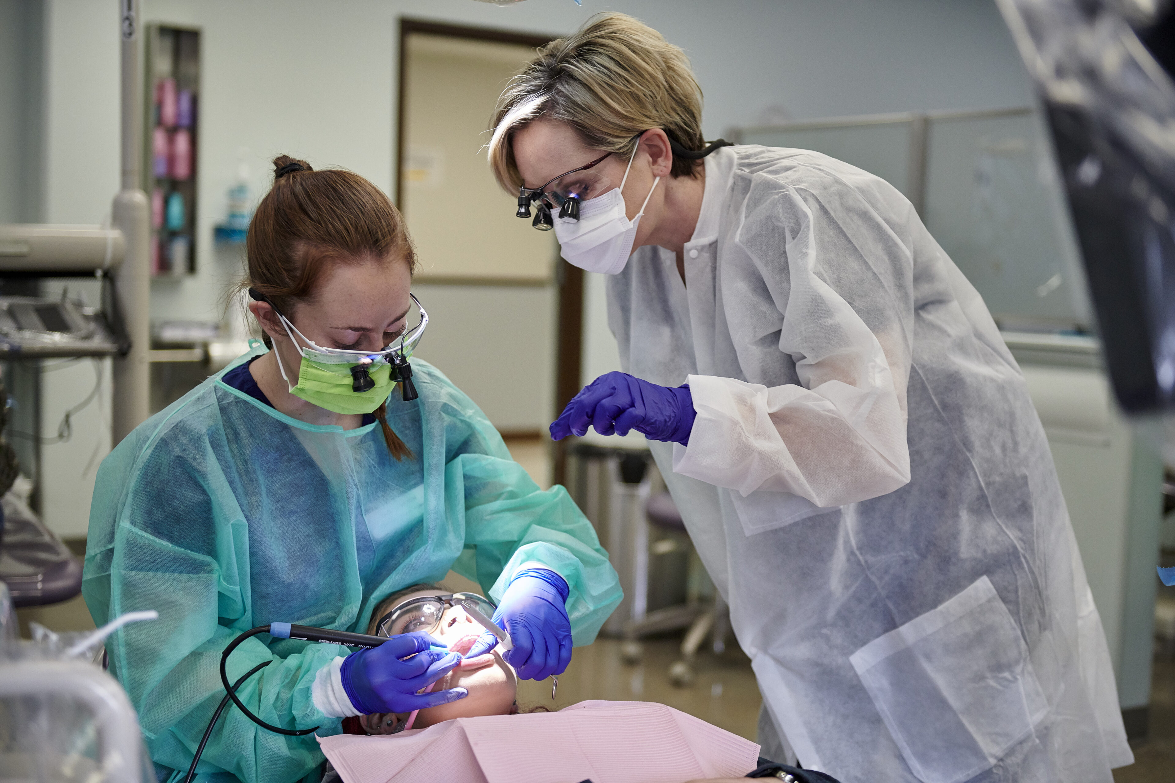 A dental professional supervises a student performing a procedure on a patient in a dental clinic, all wearing protective gear and masks.