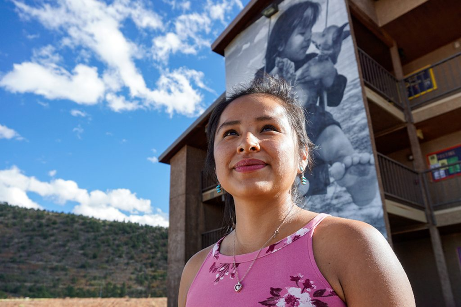 A woman in a pink top stands outside a building with a large mural of a child and dog, under a partly cloudy blue sky.