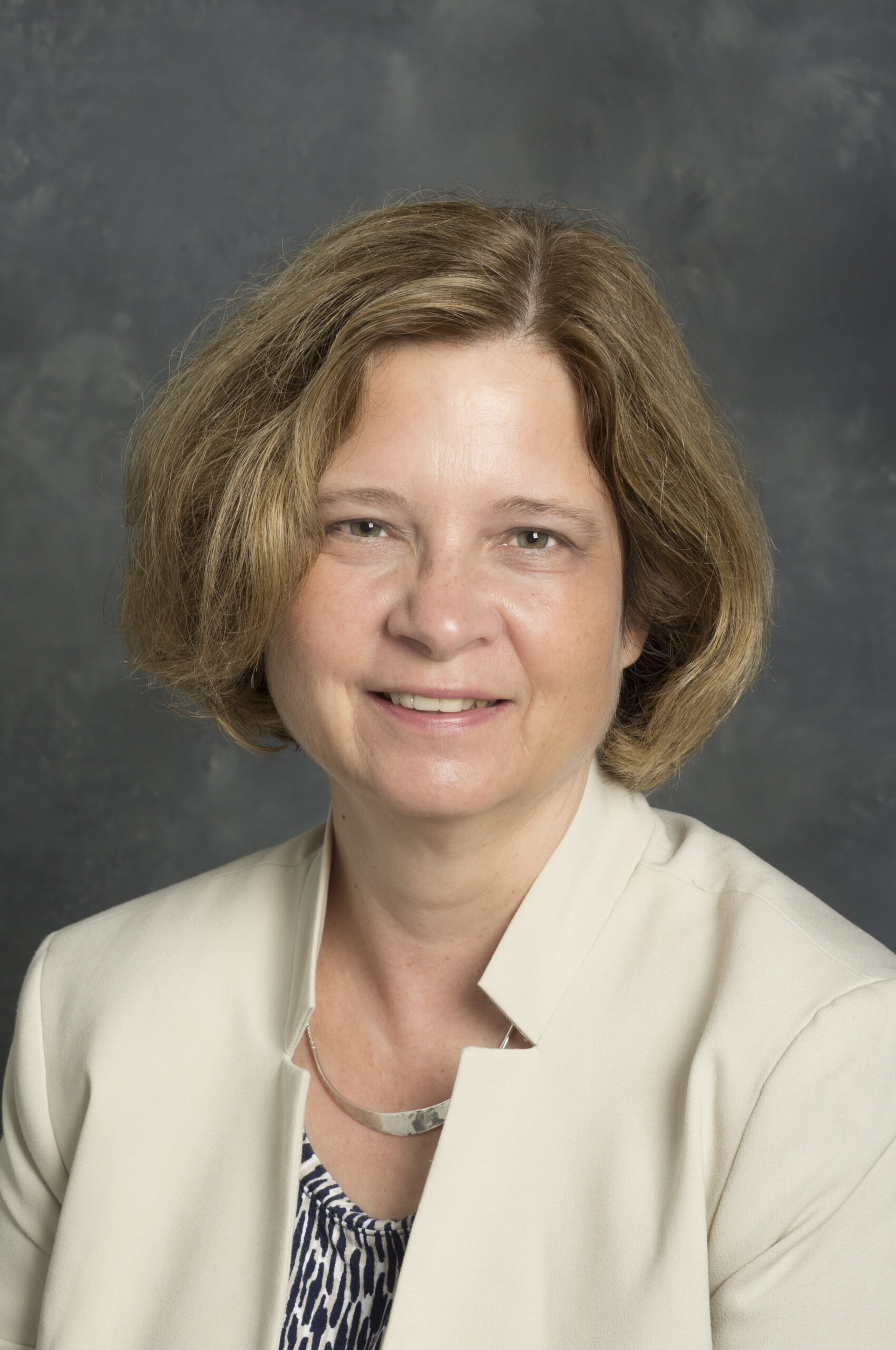 A woman with short light brown hair wearing a beige blazer and patterned blouse, posed in front of a gray background.