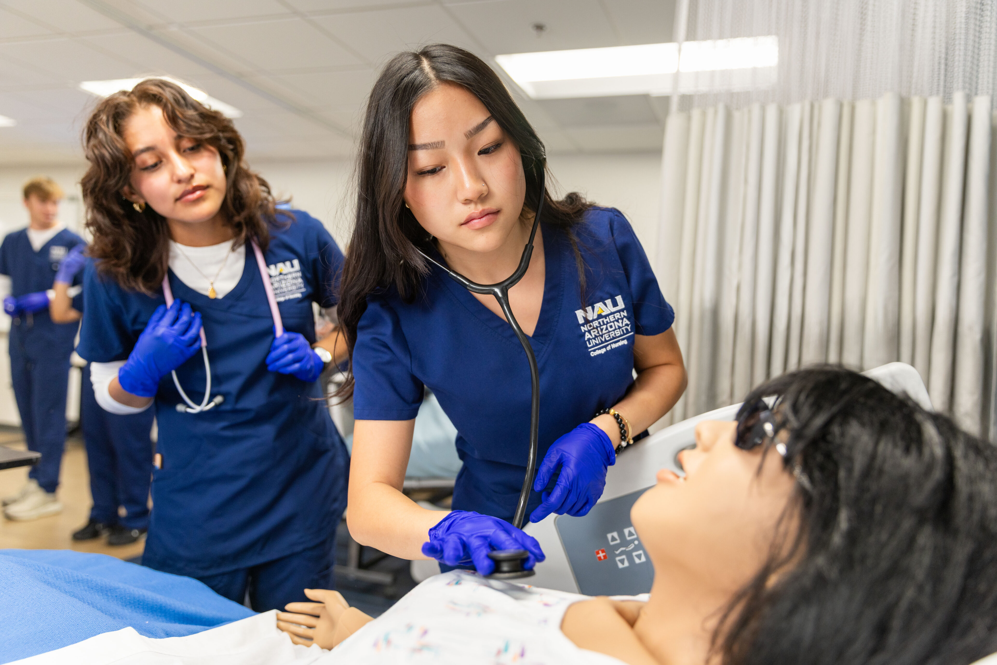 Two people in blue medical uniforms examining a patient lying on a hospital bed. The examinees appear to be medical professionals performing a medical procedure. One person has long dark hair, while the other has curly hair.