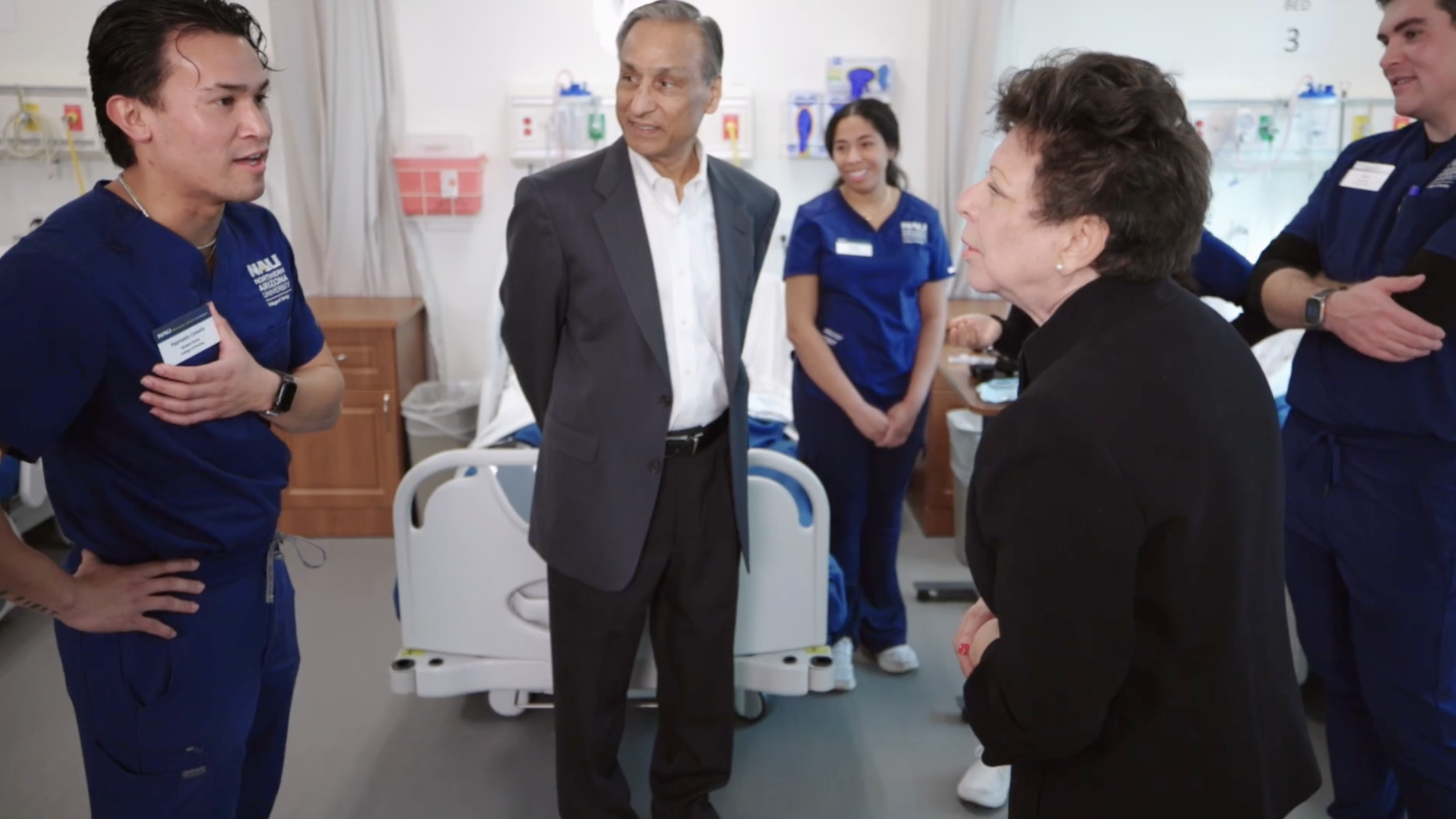 Maria and Steve Sanghi talk to a male nursing student wearing navy blue scrubs standing in front of a hospital bed.