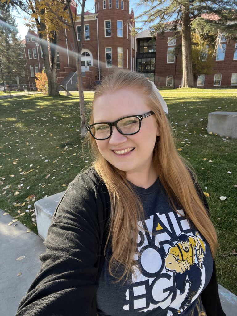 Jocelyn Whiteneck stands on NAU's north campus with Old Main in the background and green grass sprinkled with gold leaves.