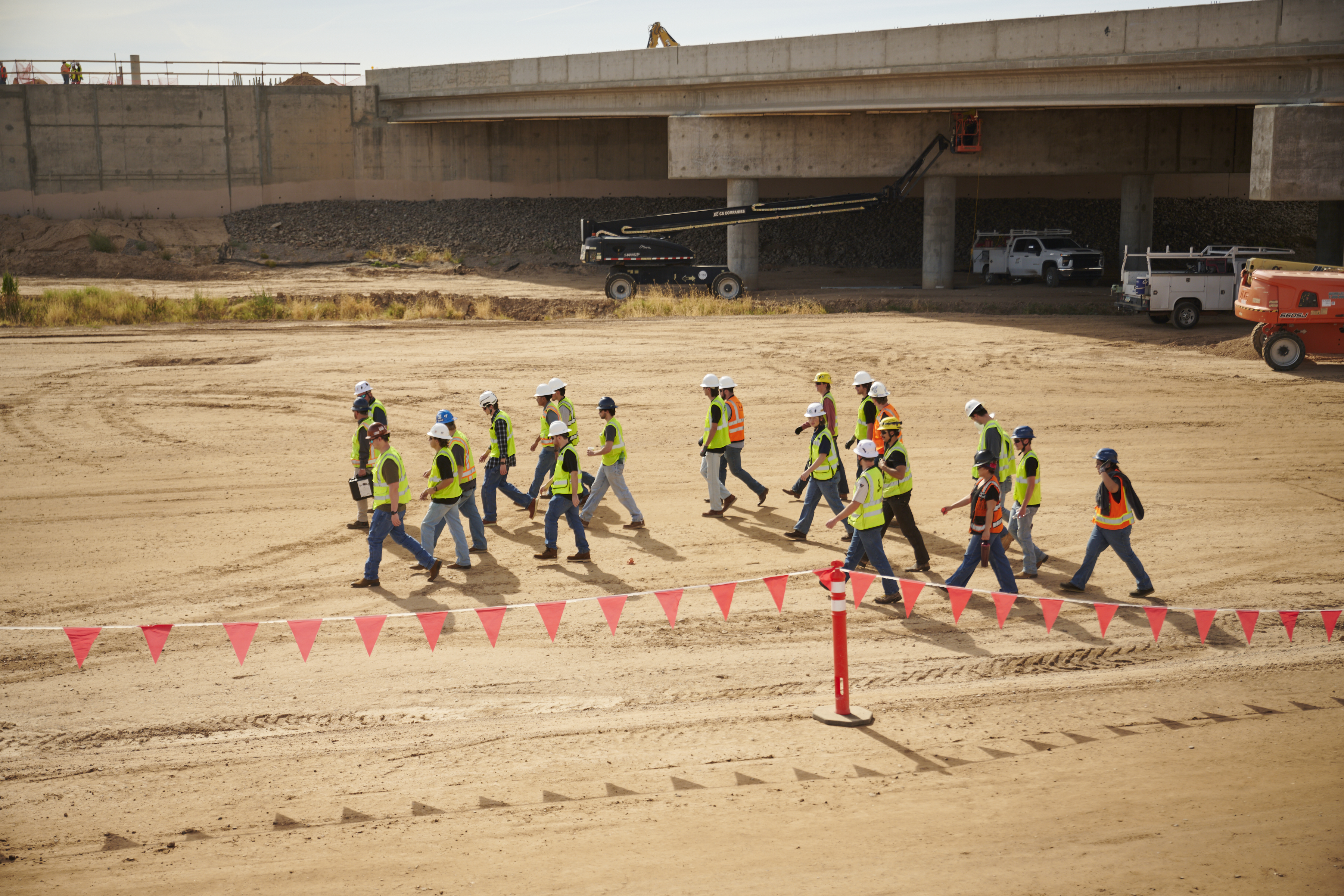 Endowed professor Kai Kaoni leads NAU students walking on dirt beneath large concrete highway bridge on construction site wearing yellow vests and white and blue hardhats