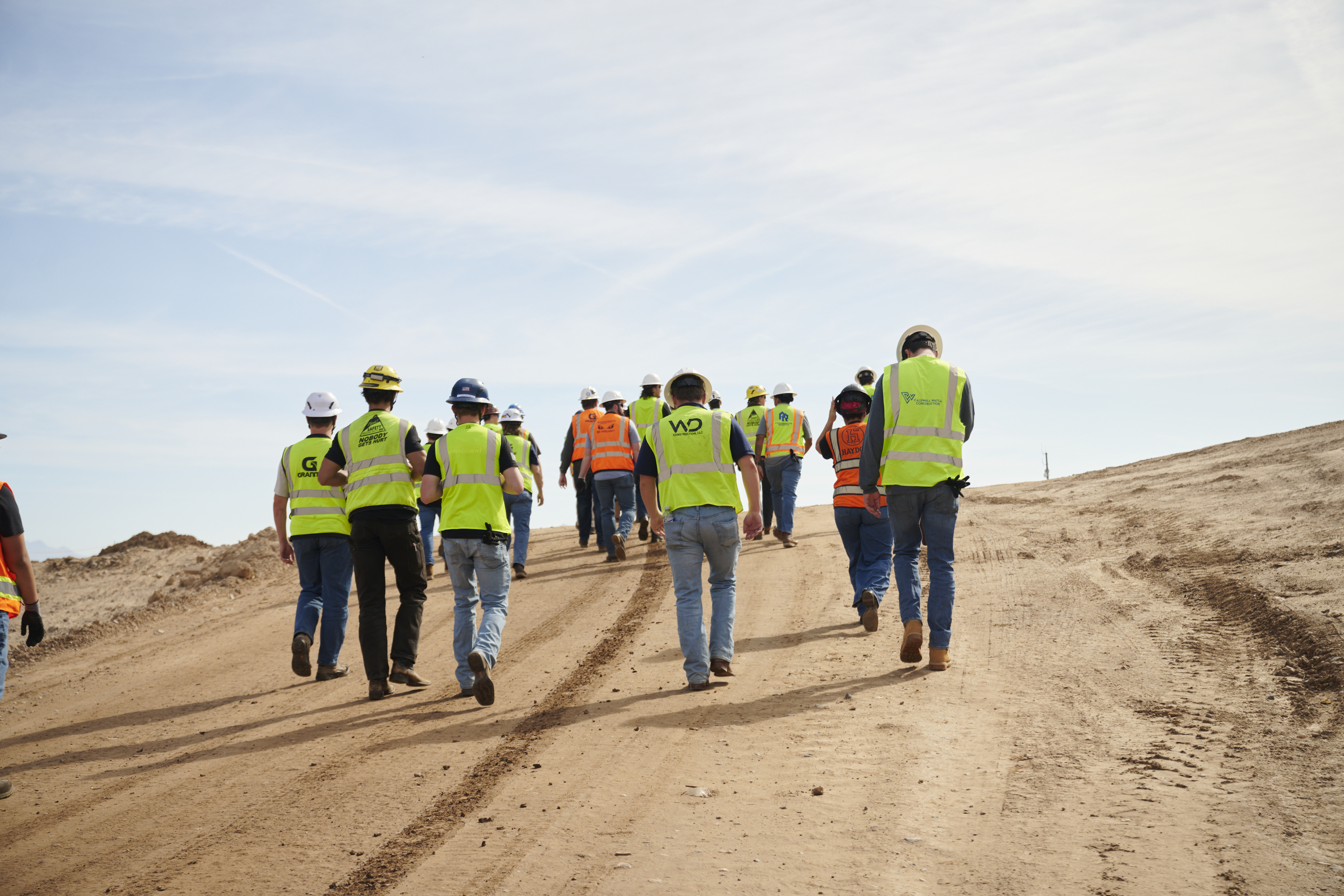 Group of a dozen students walk up a dirt hill with light blue sky in the background with their backs to the camera wearing bright yellow vests and hard hats.