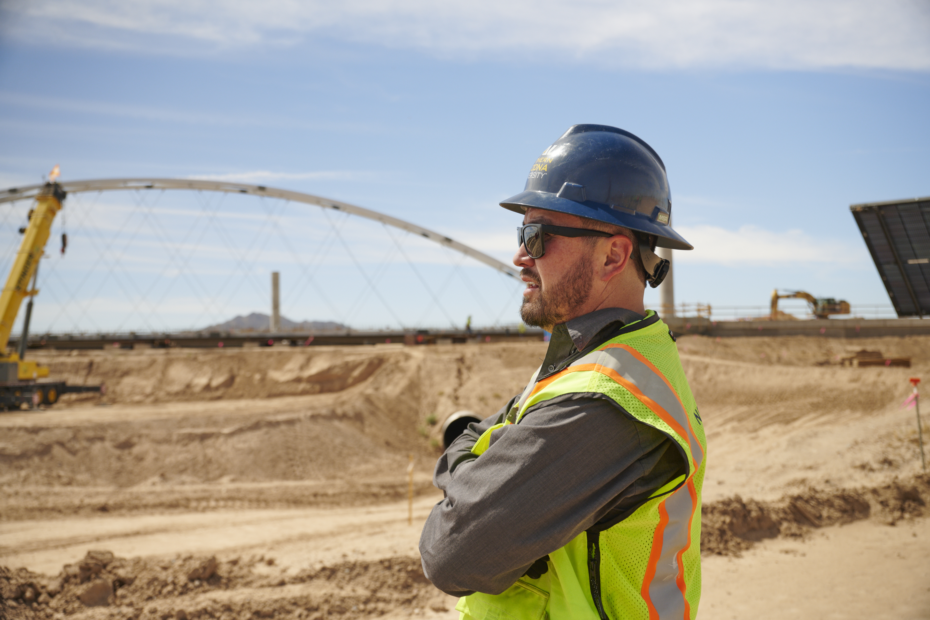 Kai Kaoni NAU Construction Management endowed professor stands on a construction site looking to the left with his arms folded over his chest wearing a blue NAU hardhat