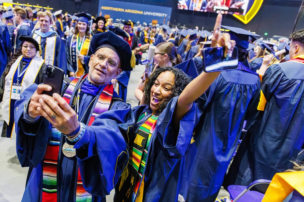 Two graduates in blue robes and mortarboards take a selfie at Northern Arizona University commencement ceremony, wearing colorful academic stoles and medals. The arena is filled with other graduates in the background.