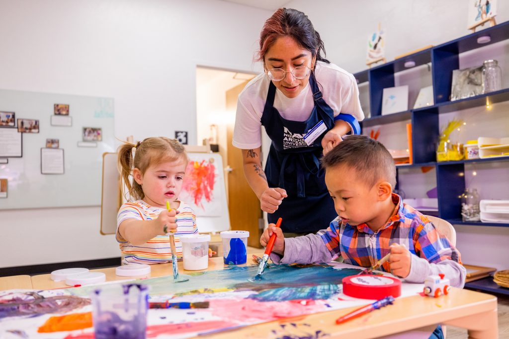 Two young children, a boy and a girl, sitting at a table with an adult woman. The children are painting or doing art activities, and the woman appears to be assisting them. The table is cluttered with art supplies and materials. The room has shelving and decorated walls, indicating it is likely an art or classroom setting.